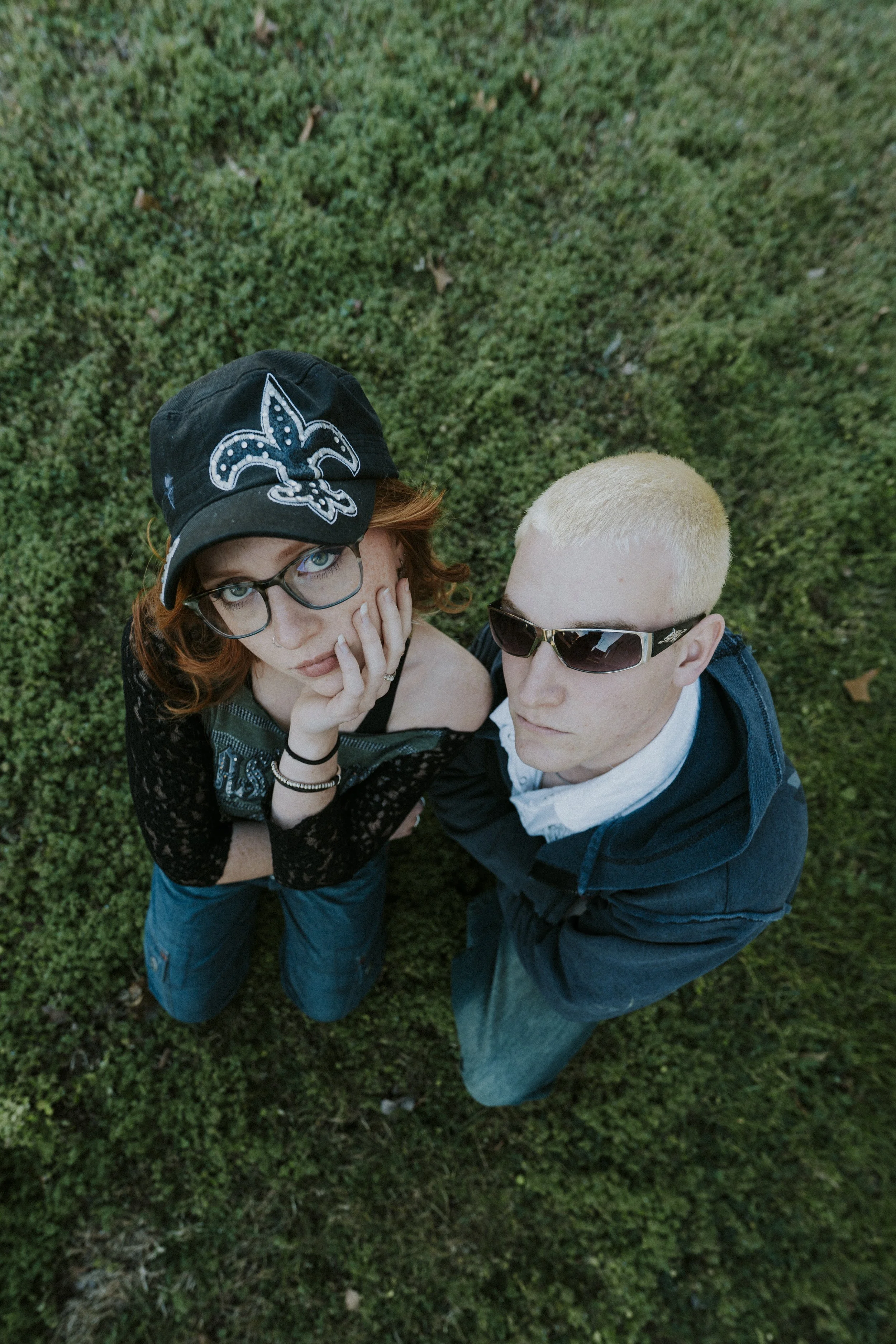 A woman with glasses and a black cap with a fleur-de-lis symbol, and a man with blond hair and sunglasses, standing on grass and looking up at the camera.