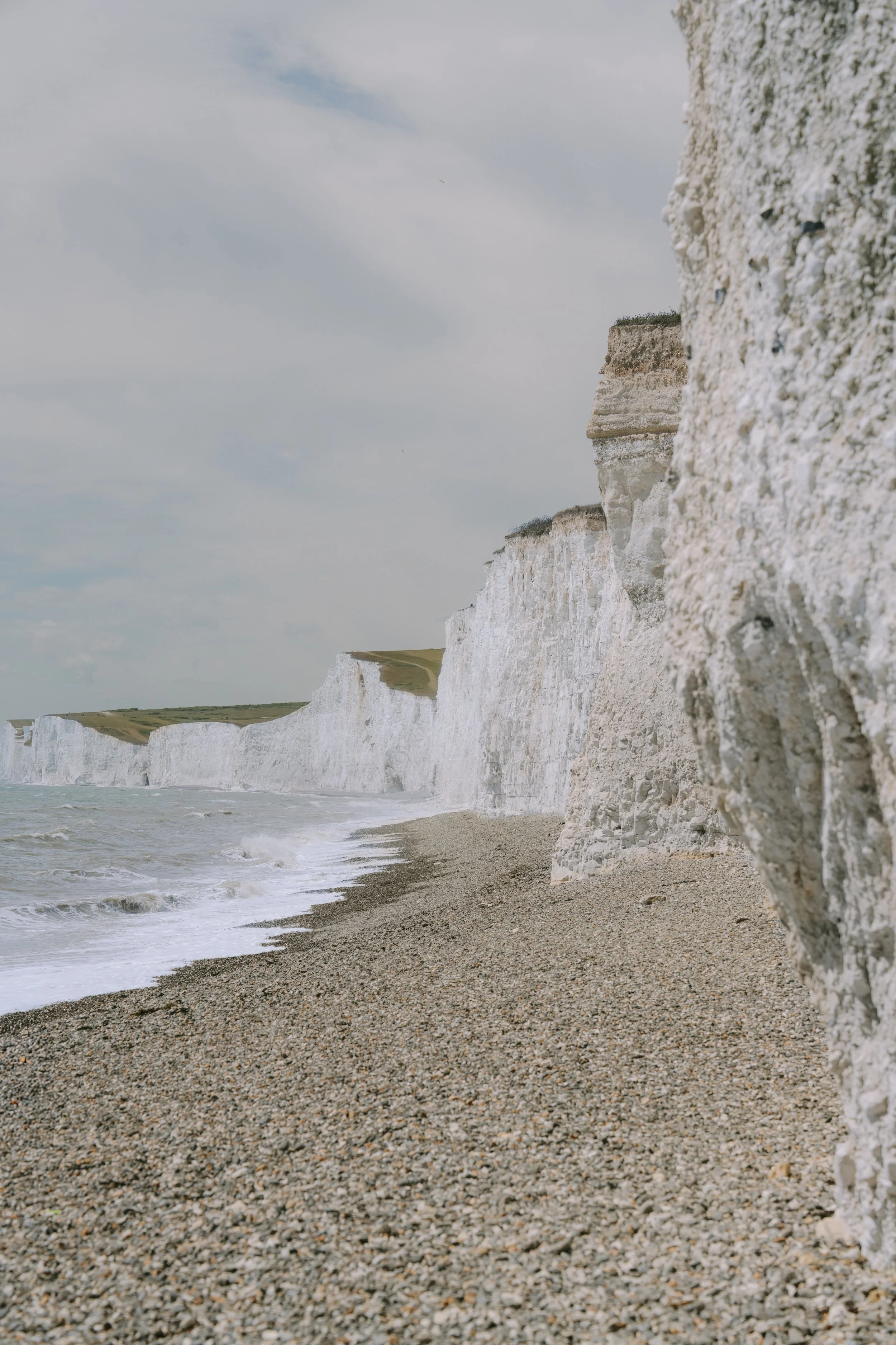 White chalk cliffs along a pebble beach with ocean waves and a cloudy sky.