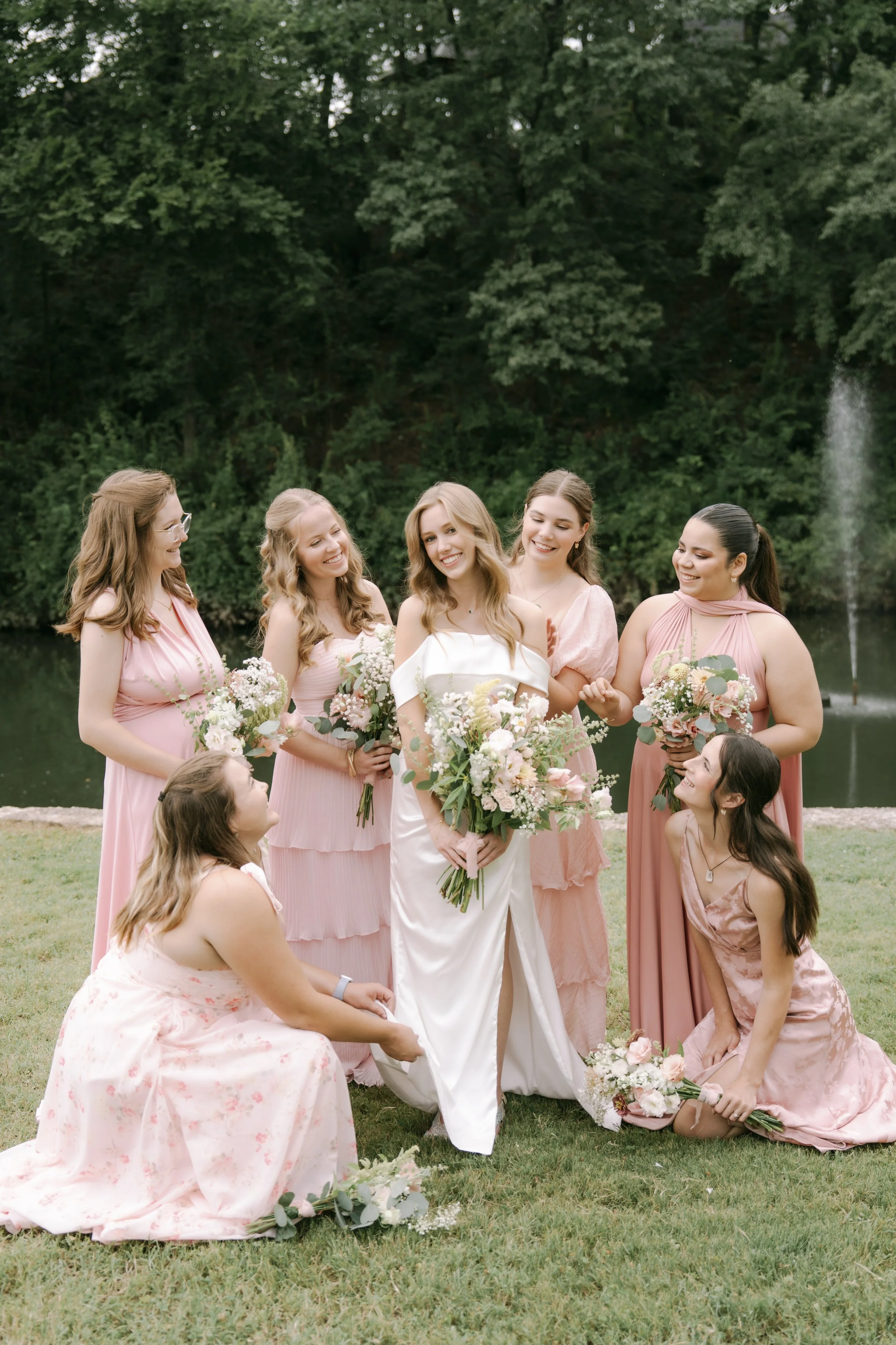 Group of women, dressed in pink and white, celebrating outdoors by a lake with a fountain, holding bouquets, smiling, during a wedding or special occasion.