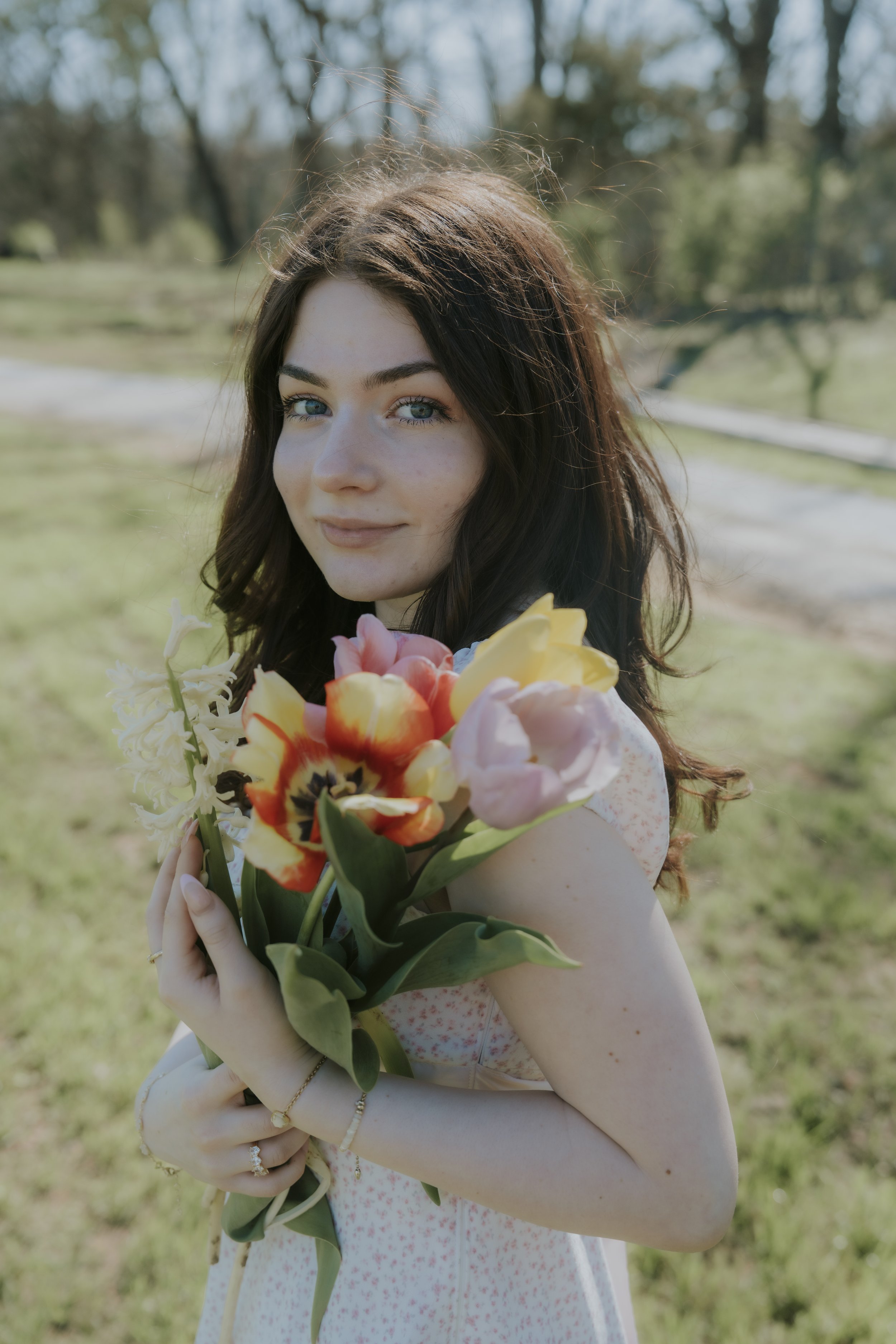 A young woman with brown hair and blue eyes holding a bouquet of colorful tulips outside on a sunny day.