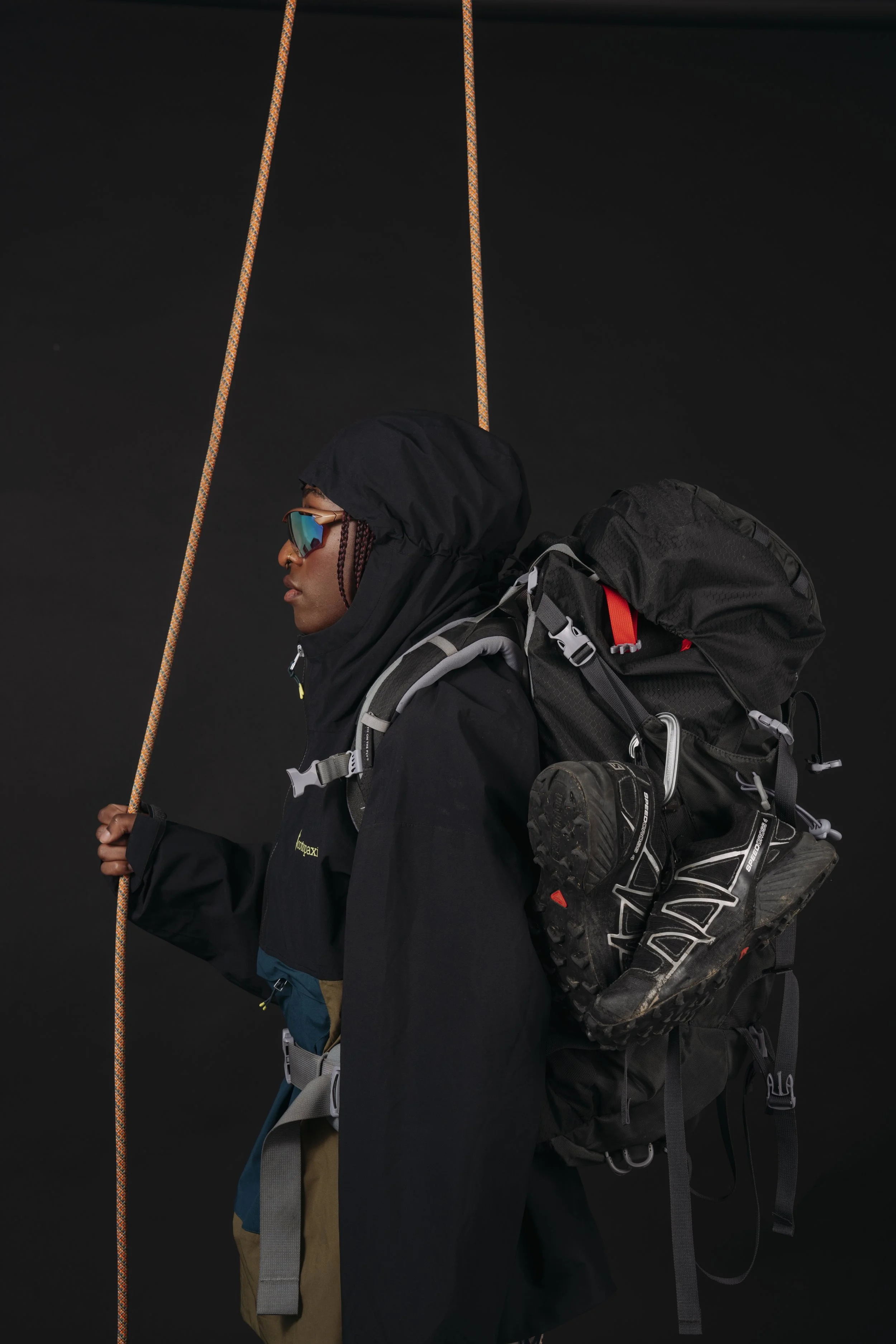 A person dressed in outdoor gear, holding a climbing or hiking rope, wearing a black jacket, tan pants, sunglasses, and carrying a large black backpack with hiking boots attached, against a dark background.