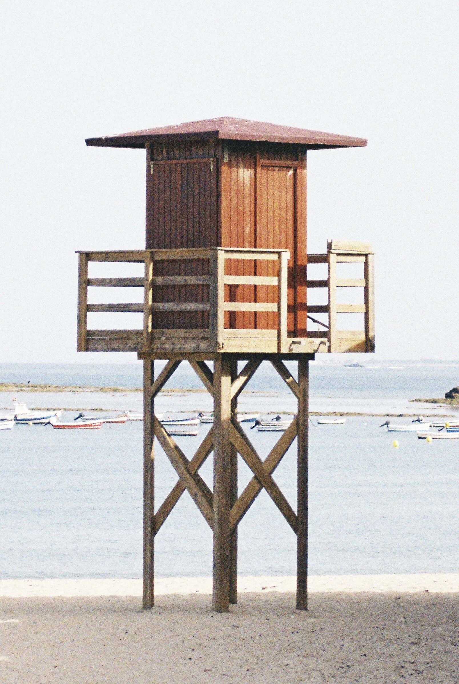 A small wooden watchtower on stilts at the beach with boats on the water in the background.