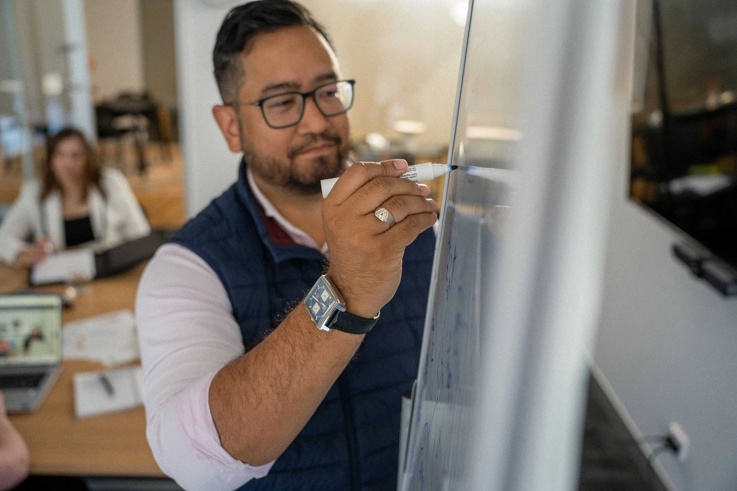 A man wearing glasses writing on a whiteboard in a meeting room, with a woman seated at a table in the background.