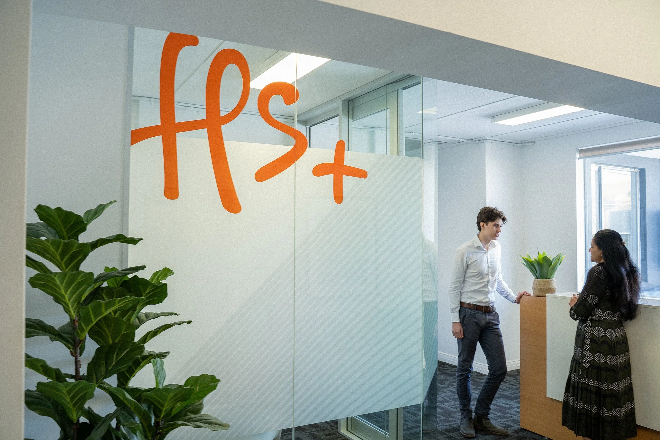 Two people, a man and a woman, are talking in an office reception area near a wooden desk. There are green potted plants and a glass wall with the orange HSPlus logo on it.