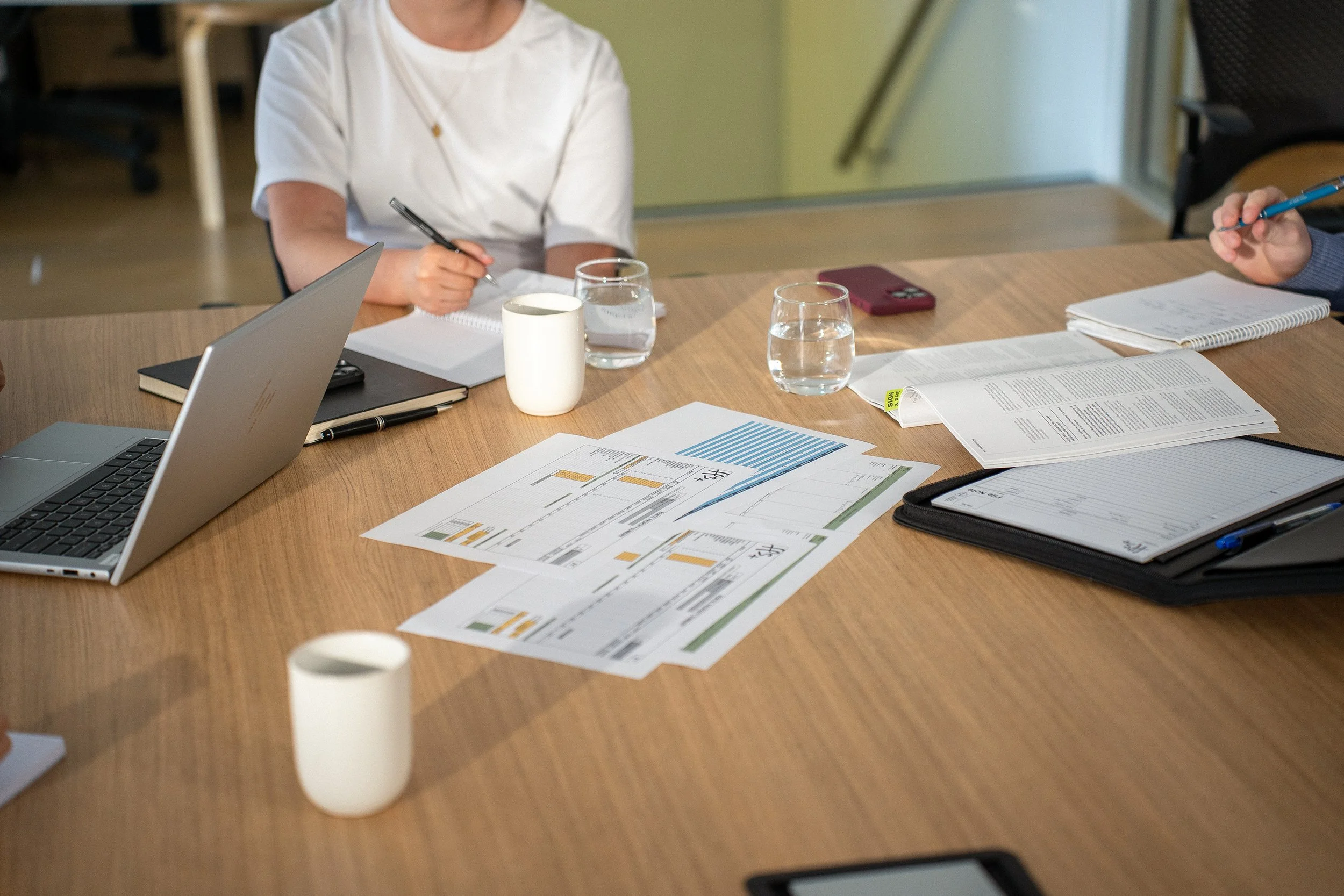 A meeting table with documents, notebooks, a laptop, glasses of water, and a person's hand holding a pen, in a professional setting.