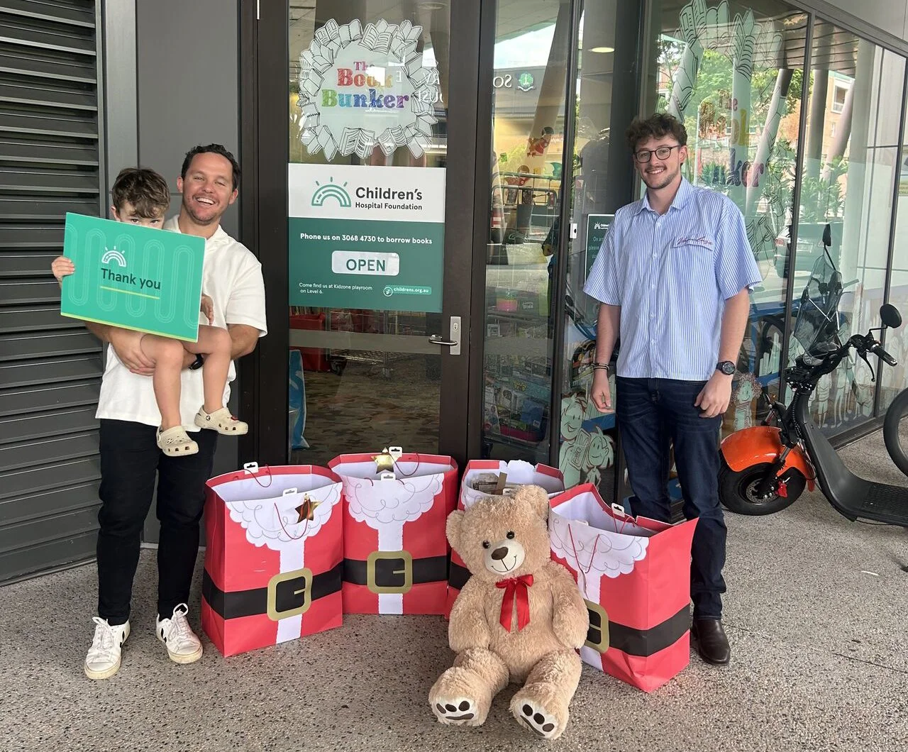 Two men and a young child standing outside the Children's Hospital Foundation building, smiling for the camera. The man on the left is holding the child, who is holding a green sign that says 'Thank you.' There are large Santa Claus-themed gift bags on the ground, along with a large teddy bear in front. A small orange electric scooter is parked nearby. The window behind them displays the hospital's signage and a door with a sign that says 'Open.'