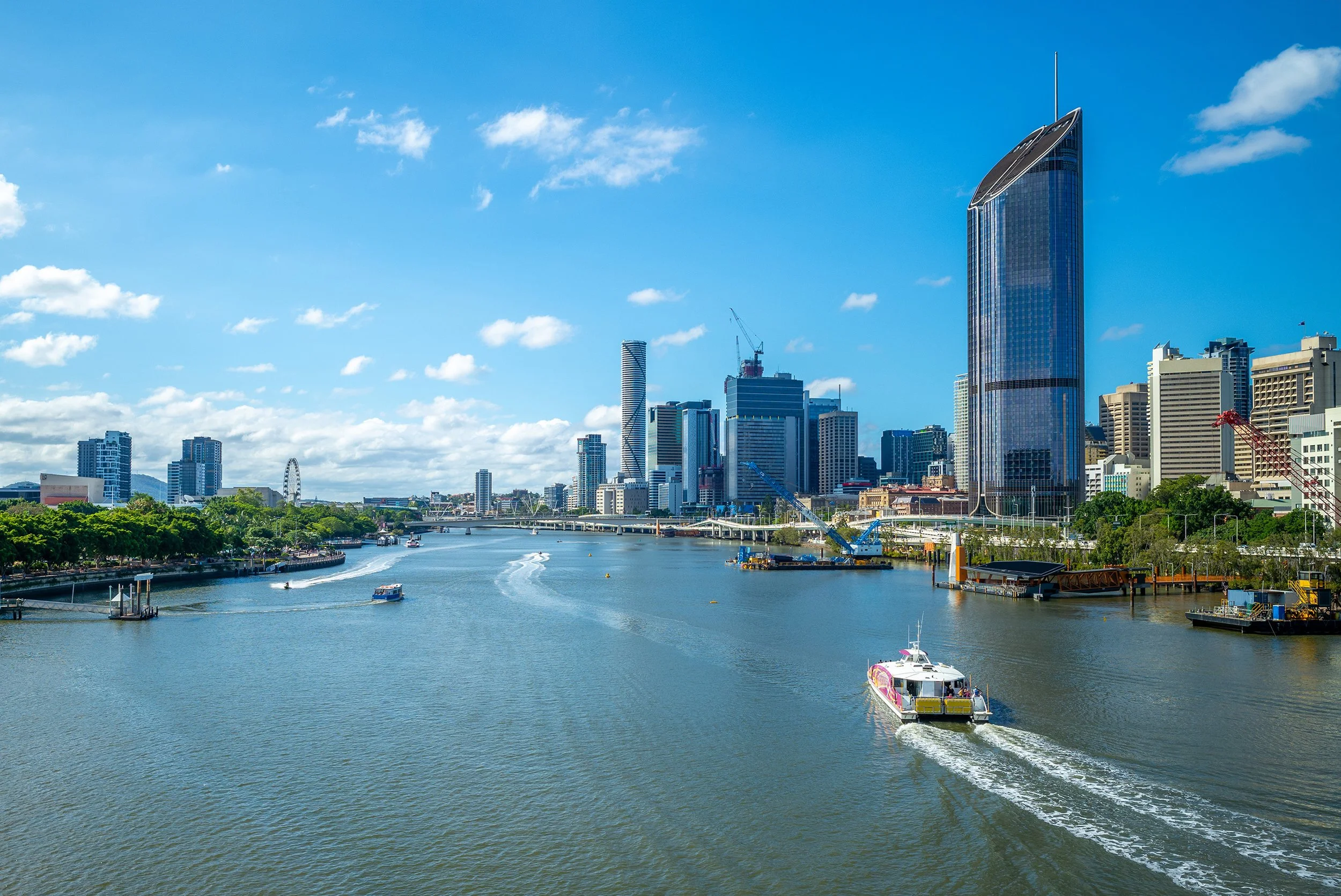 Brisbane city skyline with tall skyscrapers along a river, boats on the water, and a partly cloudy blue sky.