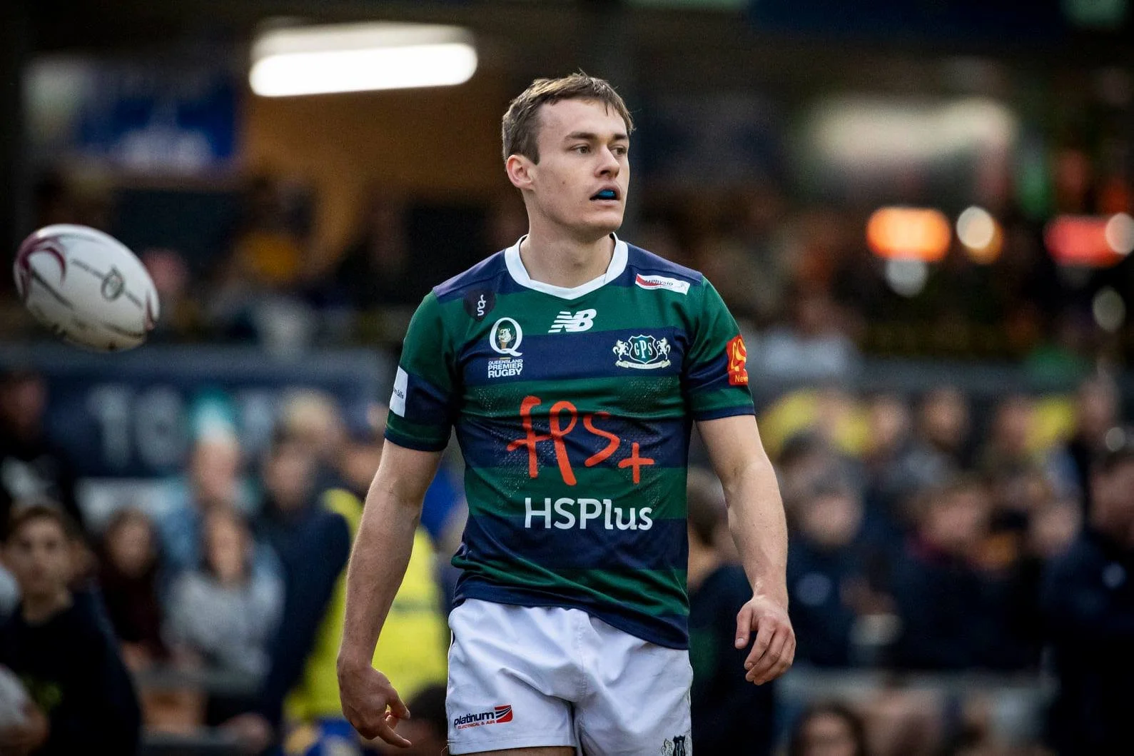 A young male rugby player with short brown hair wearing a green and blue jersey with various logos, standing on a rugby field during a game or practice, with a blurred crowd and stadium lights in the background.