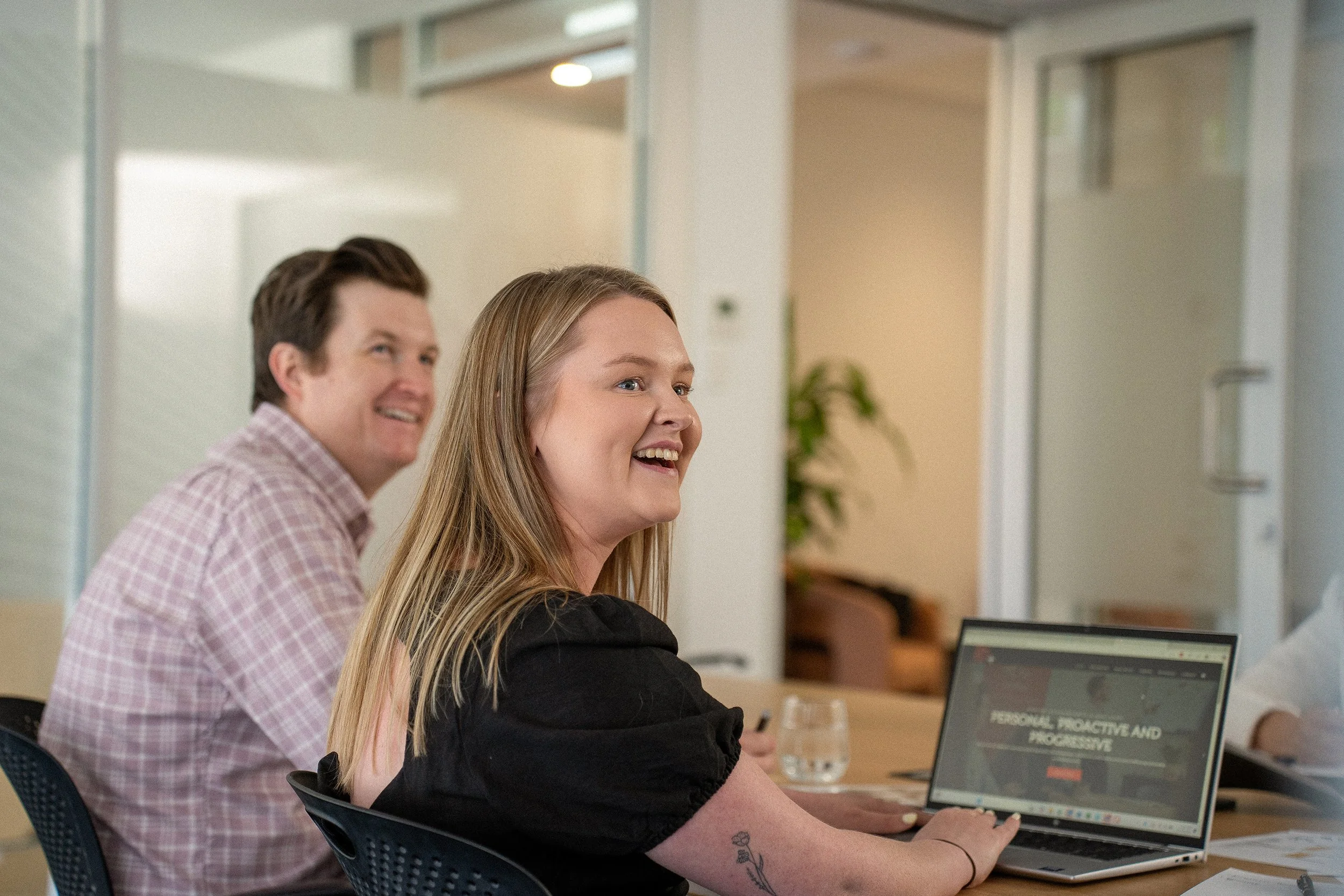 Two people, a man and a woman, sitting at a table in a modern office or conference room. The woman is working on a laptop labeled 'PERSONAL, PROACTIVE, AND PROGRESSIVE,' and the man is looking on smiling. There is a glass of water in front of the woman and a background with open doorways and a green plant.