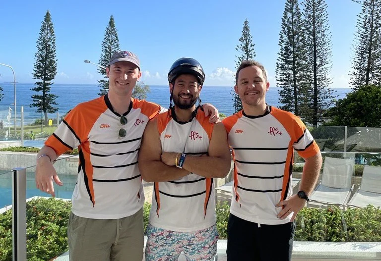 Three young men standing outdoors by a pool, with the ocean, trees, and a blue sky in the background. They are smiling and wearing matching orange and white sports shirts.