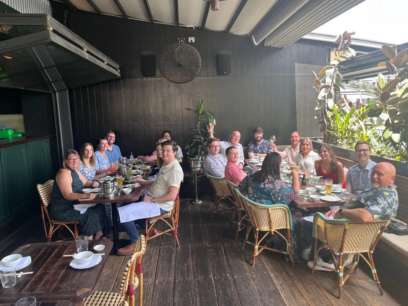 A group of people having a meal at a restaurant table outdoors near large green plants and windows.