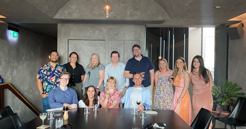 Group of 12 people gathered in a modern conference room, some seated at a table with drinks, others standing behind, smiling for a photo.