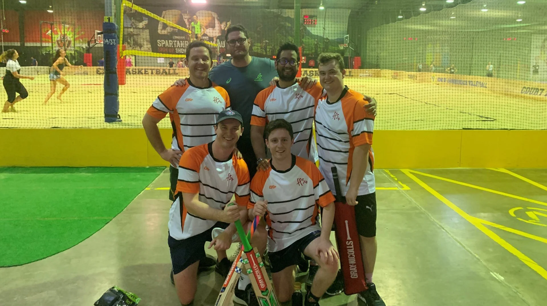 Group of seven young men in sports uniforms posing together after a match at an indoor volleyball court, with some players in action visible behind a net in the background.