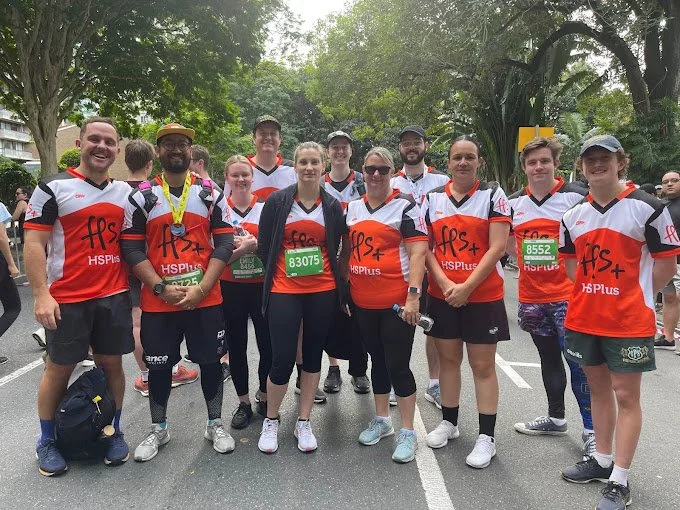 Group of people in running attire standing outdoors, smiling, with race bibs and medals, likely after a race event.