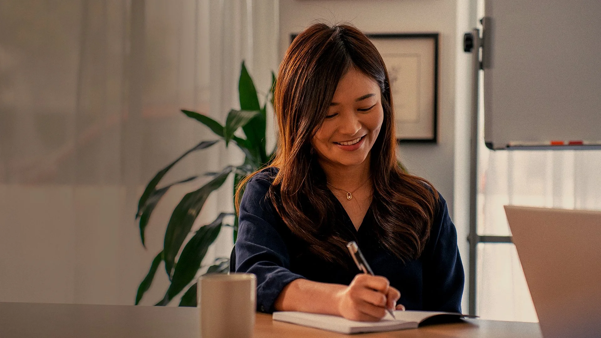 A woman with long brown hair, wearing a dark blue shirt, sitting at a desk, smiling, and writing in a notebook with a pen. There is a plant in the background and a white mug on the desk.