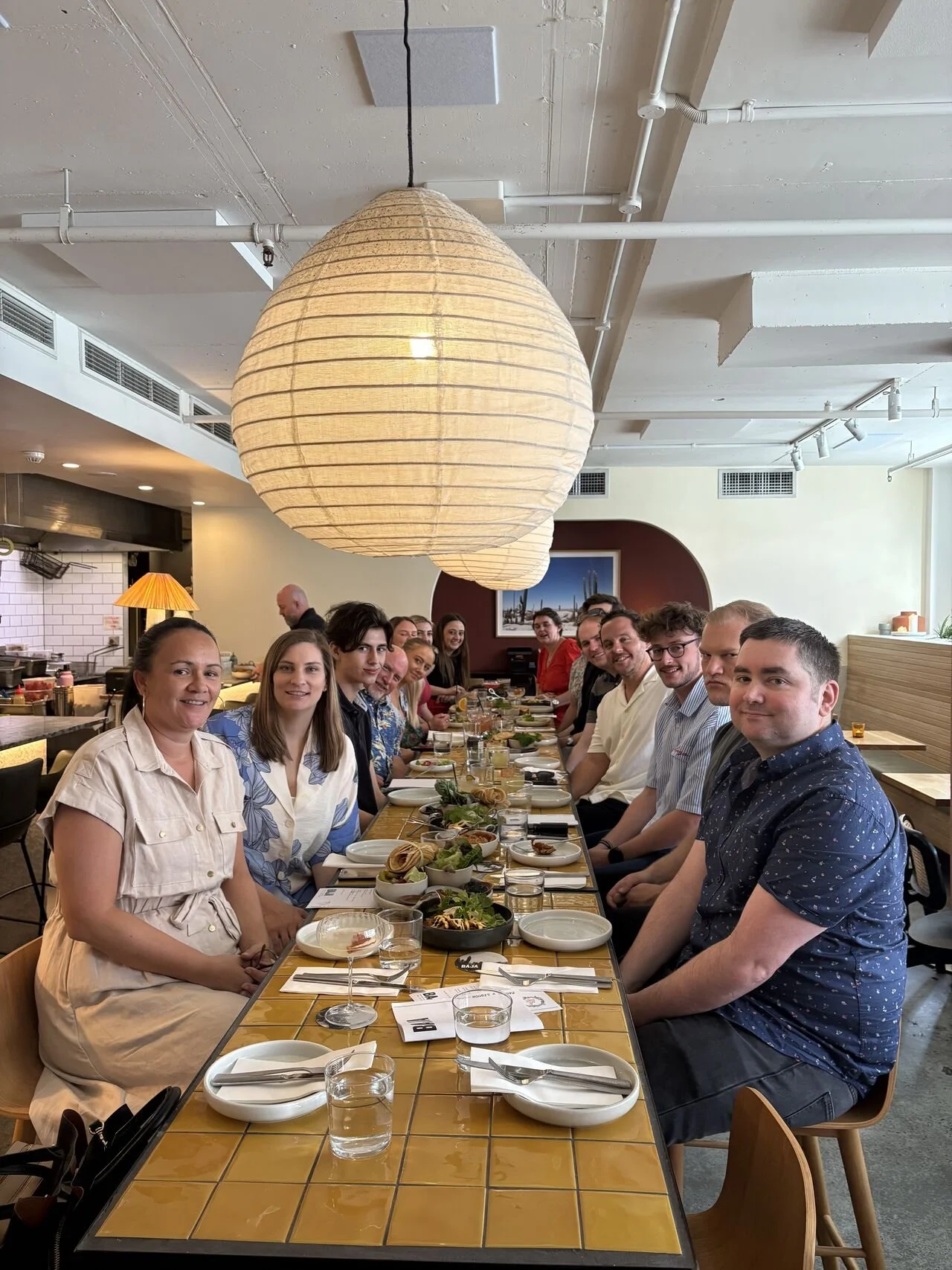 A group of people sitting at a long restaurant table, smiling, with plates of food and glasses of water, in a well-lit dining room with large paper lantern overhead.