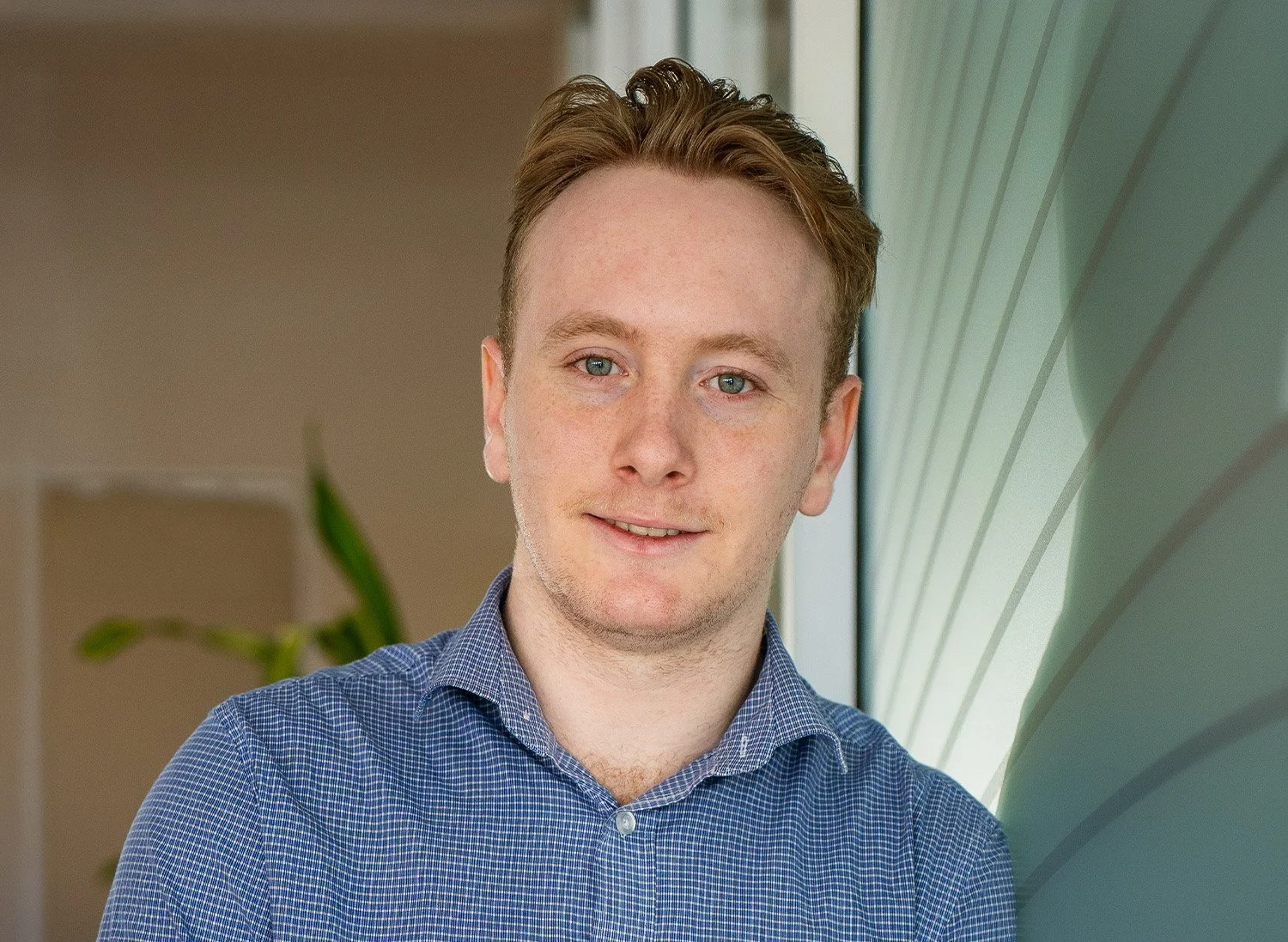 A young man with brown hair and blue eyes, wearing a blue checkered shirt, standing indoors next to a window with green curtains.