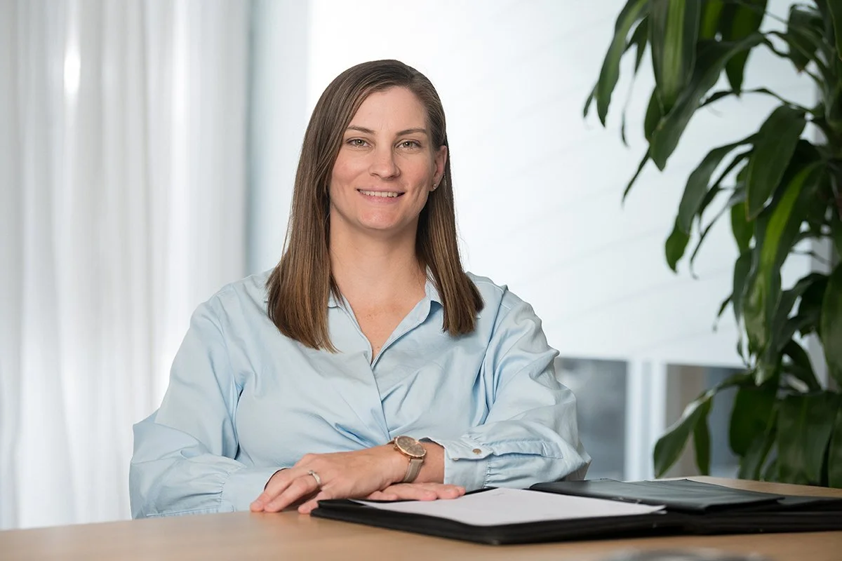 A woman with straight brown hair, wearing a light blue shirt, sitting at a desk with her arms crossed, smiling, in an office setting with a large plant to her right.