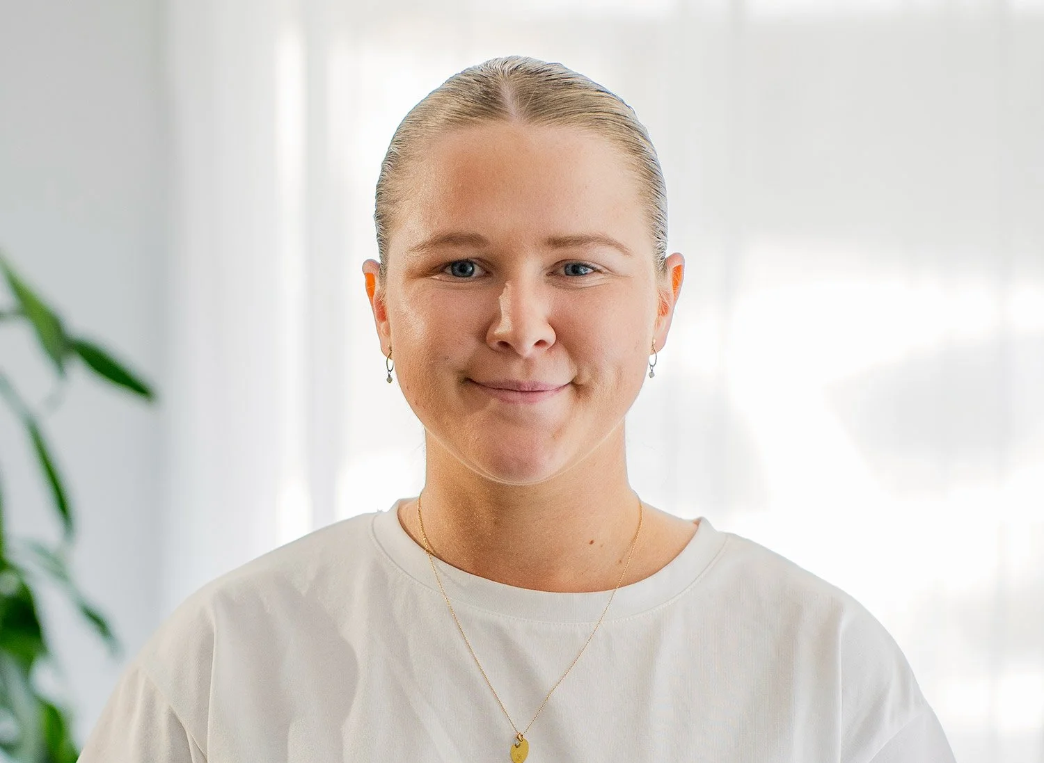 A young woman with blonde hair tied back, smiling, wearing earrings, a gold necklace with a heart-shaped pendant, and a white t-shirt in a bright room with white curtains and a green plant in the background.