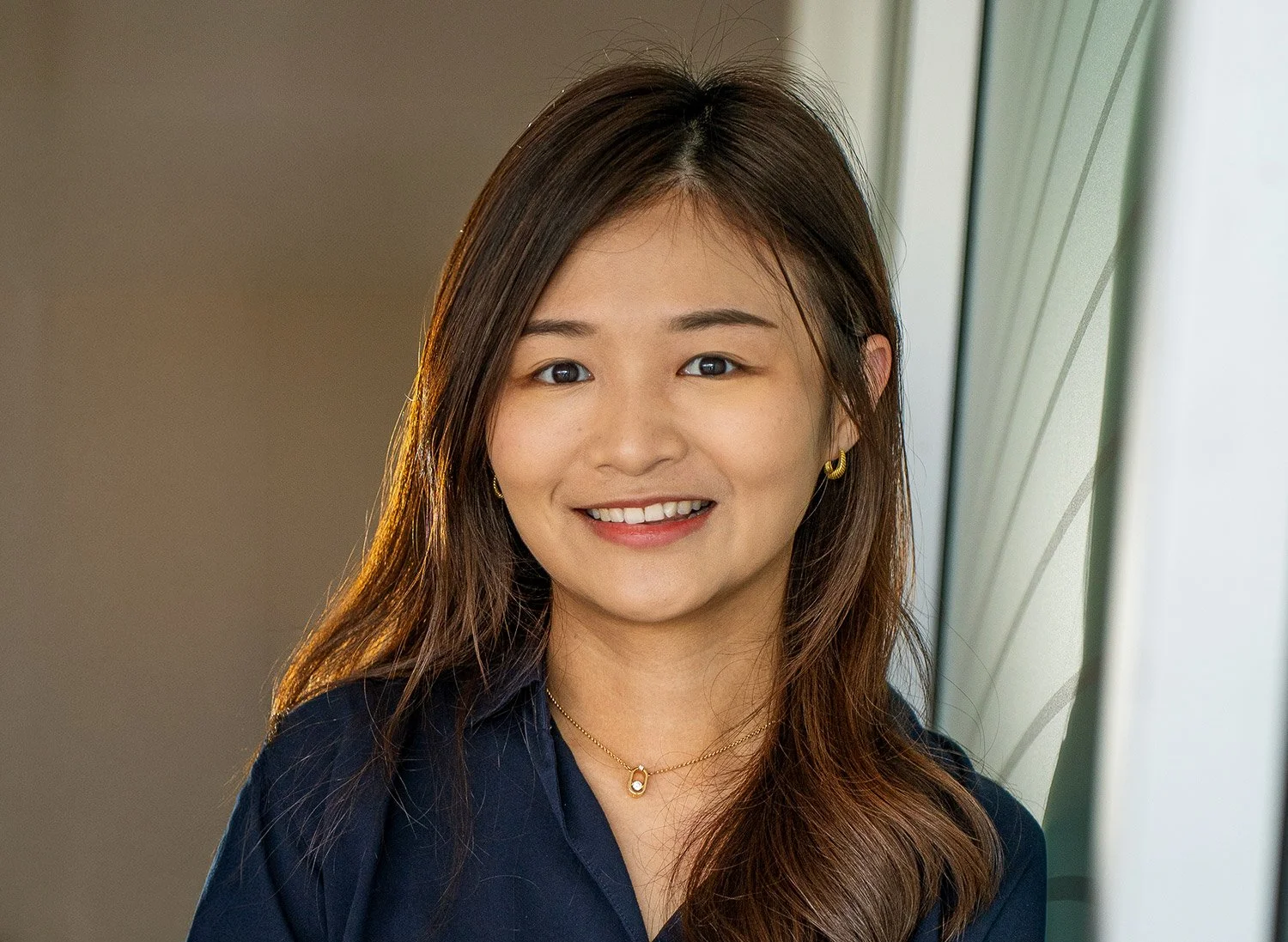 A young woman with long brown hair smiling and looking at the camera, standing near a window.