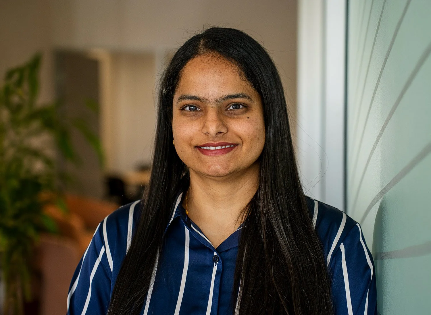 A woman with long black hair, wearing a blue and white striped shirt, smiling in an indoor office space.