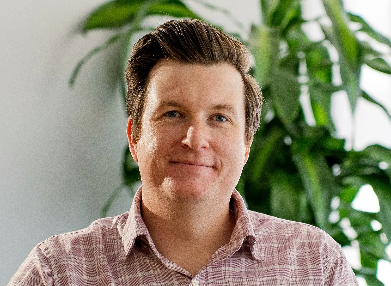 A man with brown hair and blue eyes smiling, wearing a light red checkered shirt, in front of a large green leafy plant and a white wall.
