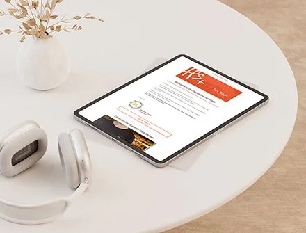 Tablet on a white desk displaying a webpage, with a white vase with dried flowers and white headphones nearby.