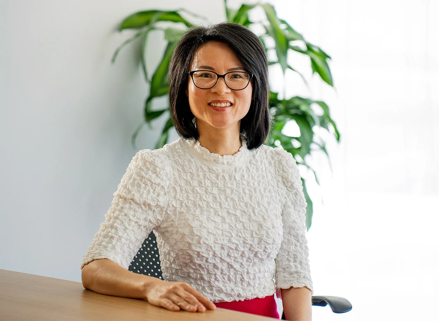 A woman with black hair and glasses smiling, sitting at a desk with a large green plant in the background.