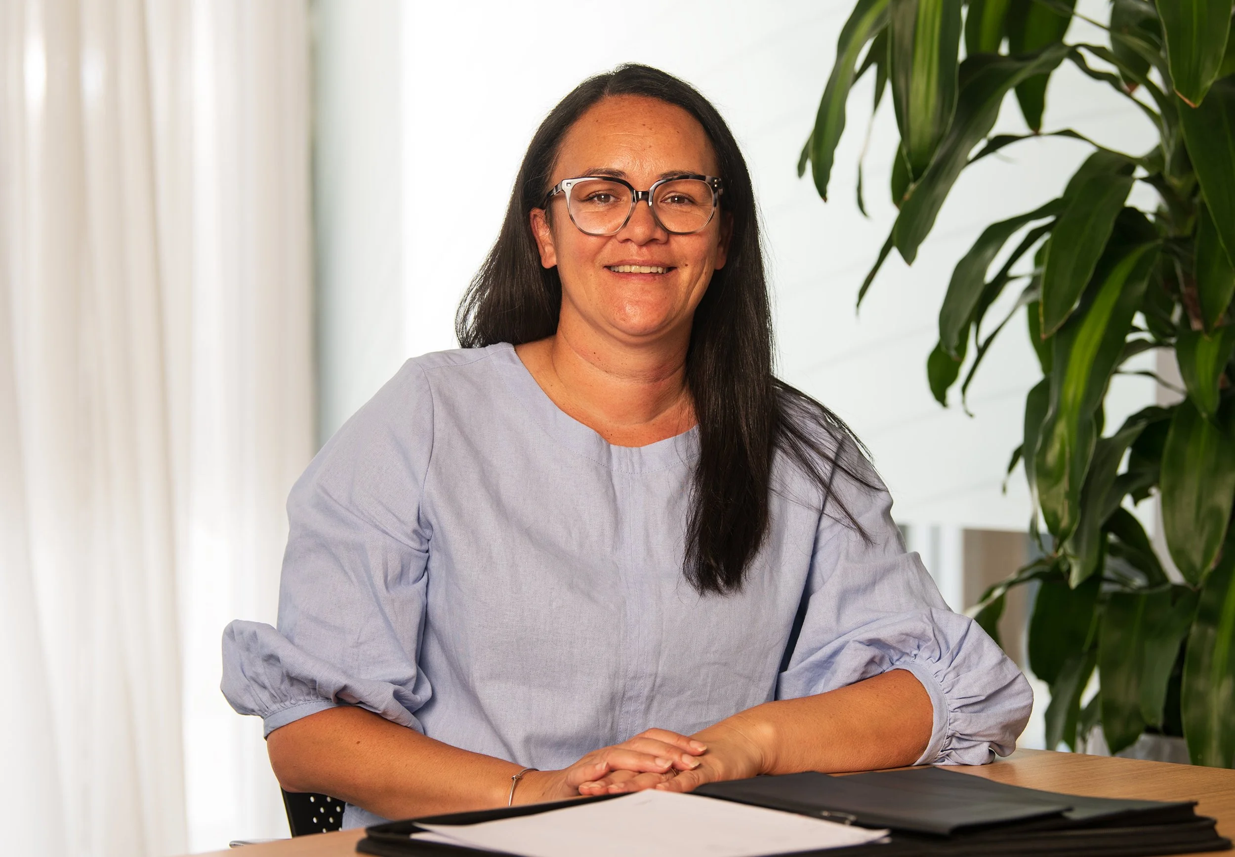 A woman with long dark hair, wearing glasses and a light purple long-sleeve shirt, sitting at a desk with papers and a laptop in front. She is smiling and has her hands clasped on the table. There is a large green houseplant behind her and a window with white curtains to her left.