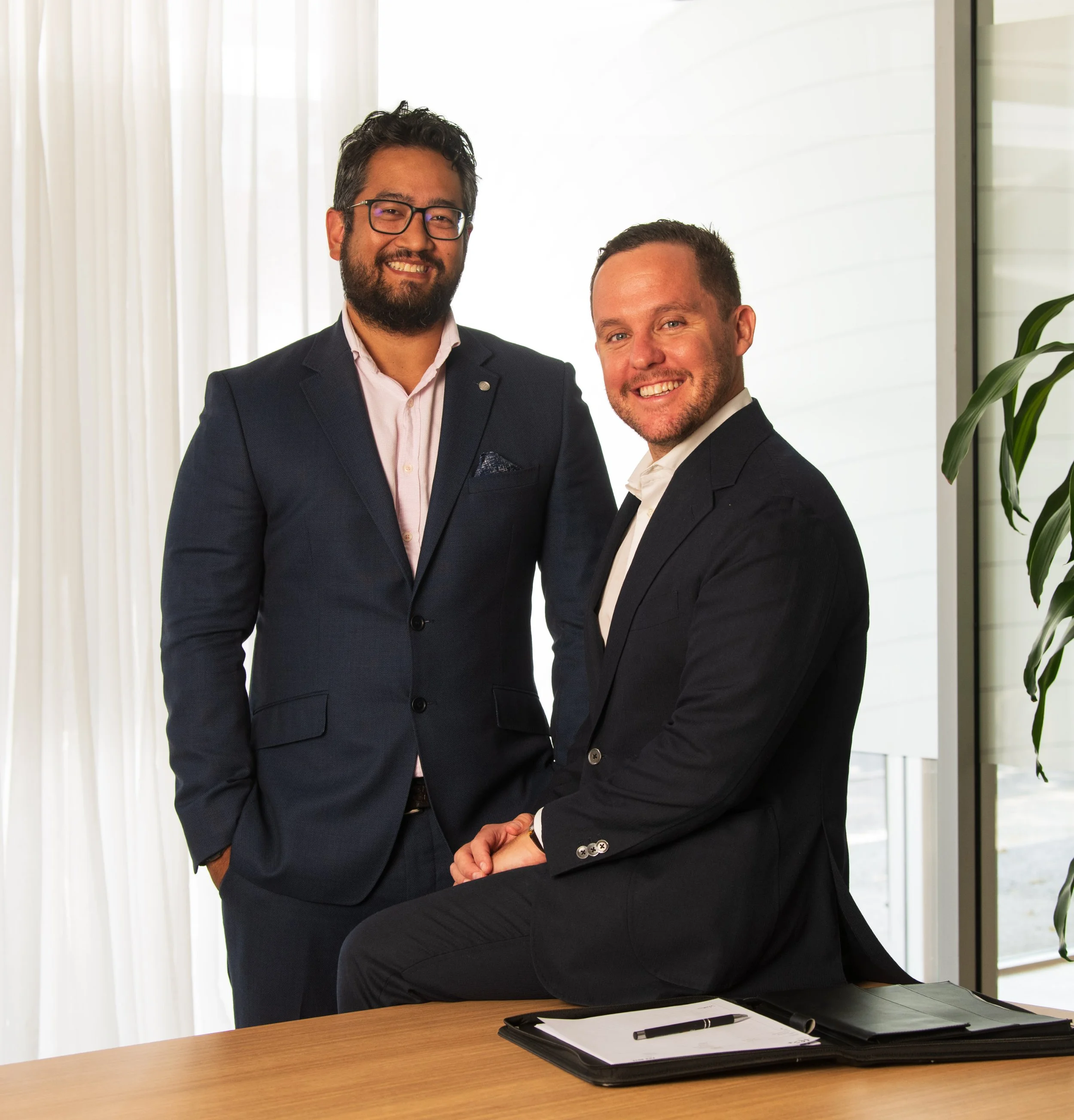 Two men in business suits smiling, standing near a table with folders, against a background of white curtains and a plant.