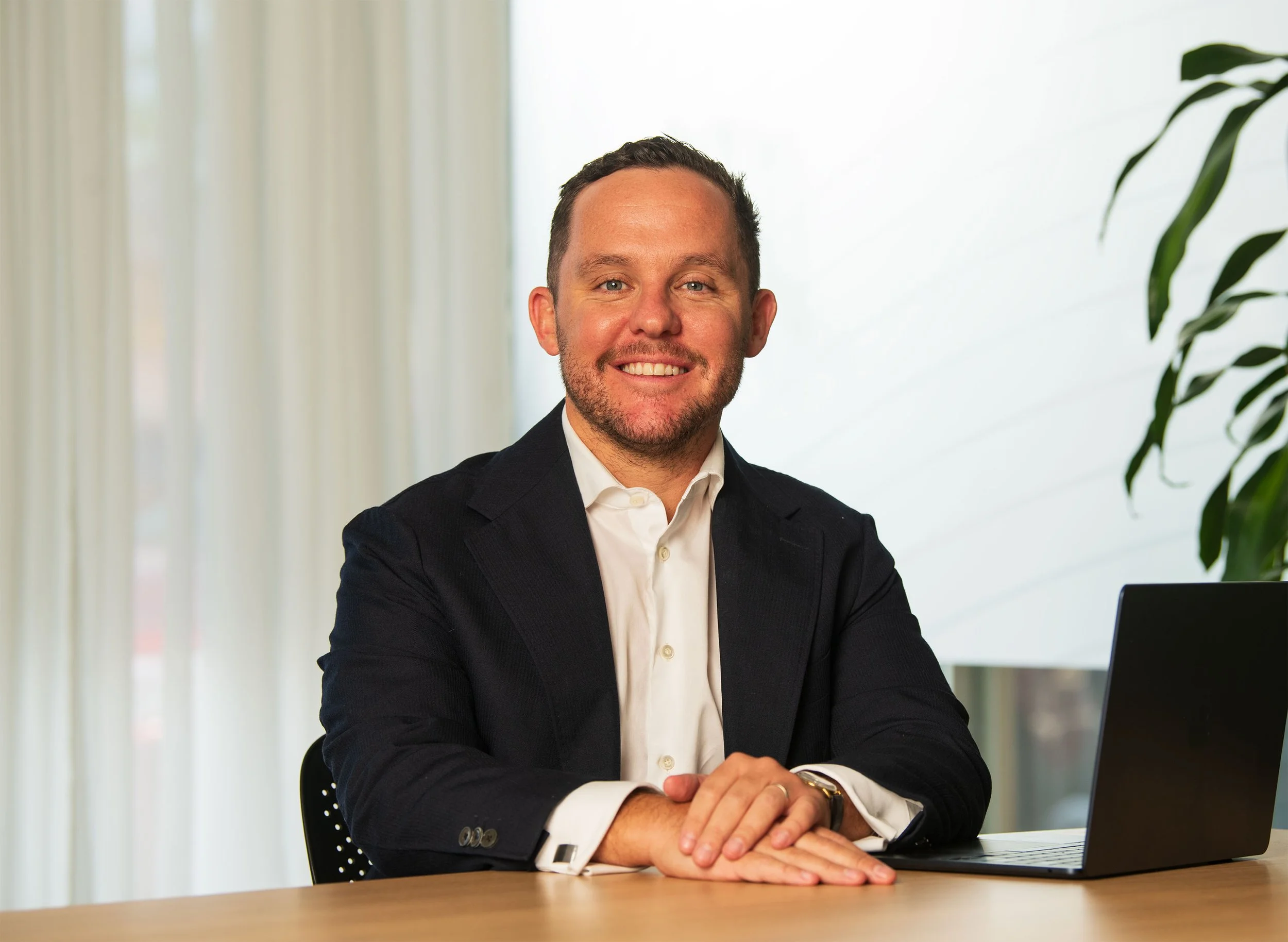 A man in a business suit smiling, sitting at a desk with a laptop in front of him, in a modern office setting.