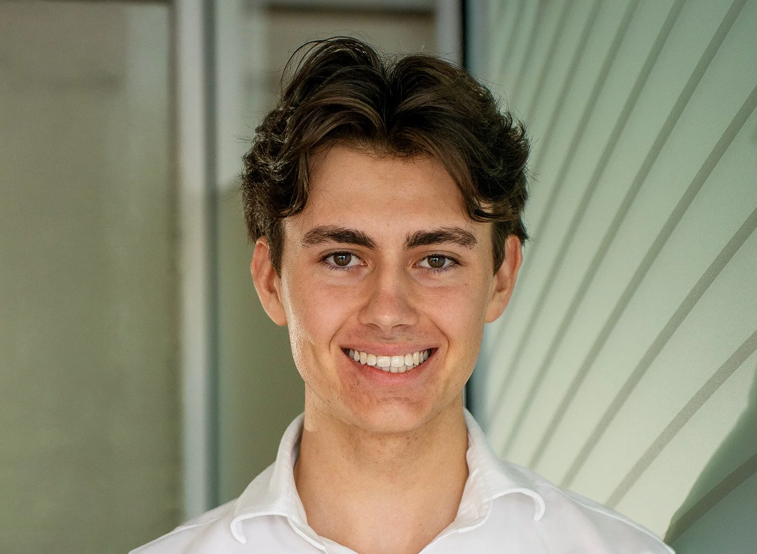 Smiling young man with brown hair wearing a white collared shirt, standing in front of a modern architectural background.