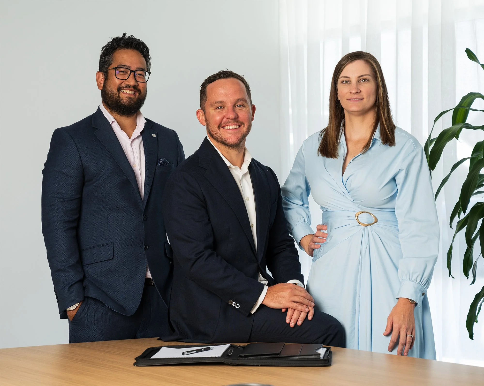 Three business professionals, two men and one woman, posing together in an office setting. The woman is standing, the men are sitting in front of a desk. There are papers, a pen, and a tablet on the desk. The background shows white curtains and a large green plant.