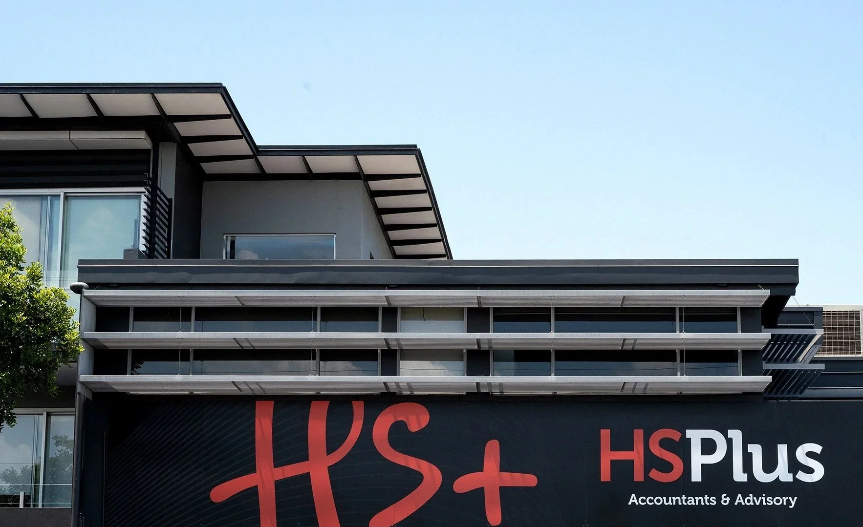 Close-up view of a modern multi-story commercial building with balconies, windows, and an office sign that reads 'HS Plus Accountants & Advisory' against a clear blue sky.