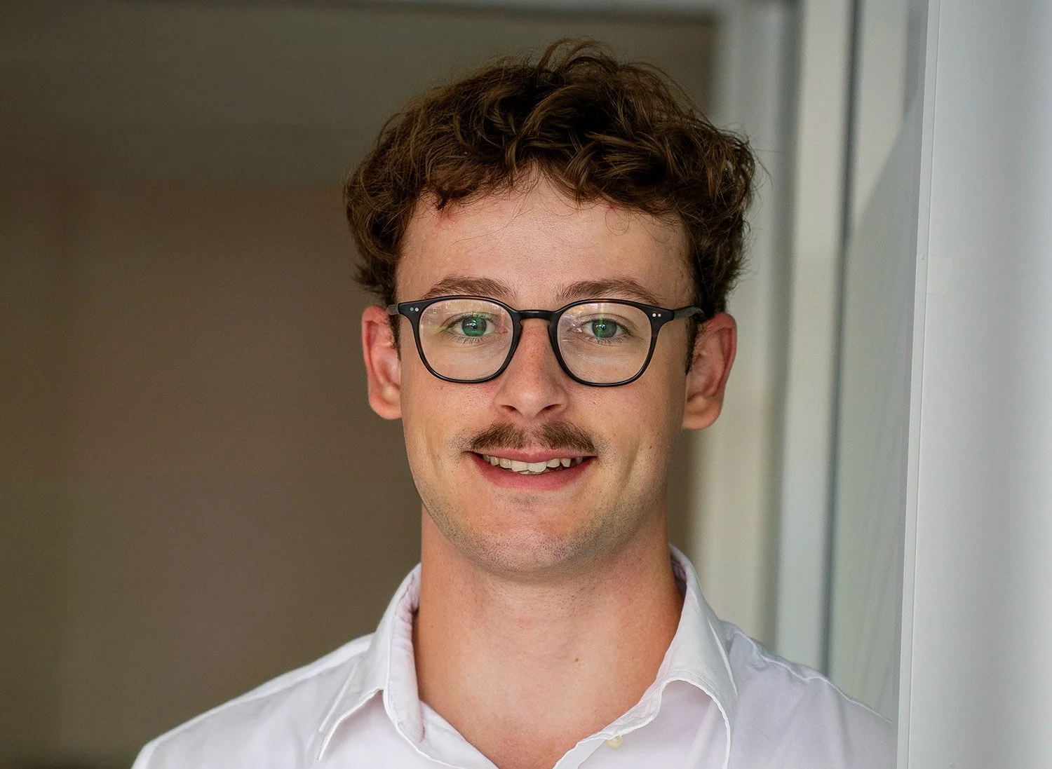 A young man with curly brown hair, wearing glasses and a white button-up shirt, smiling and looking at the camera indoors.