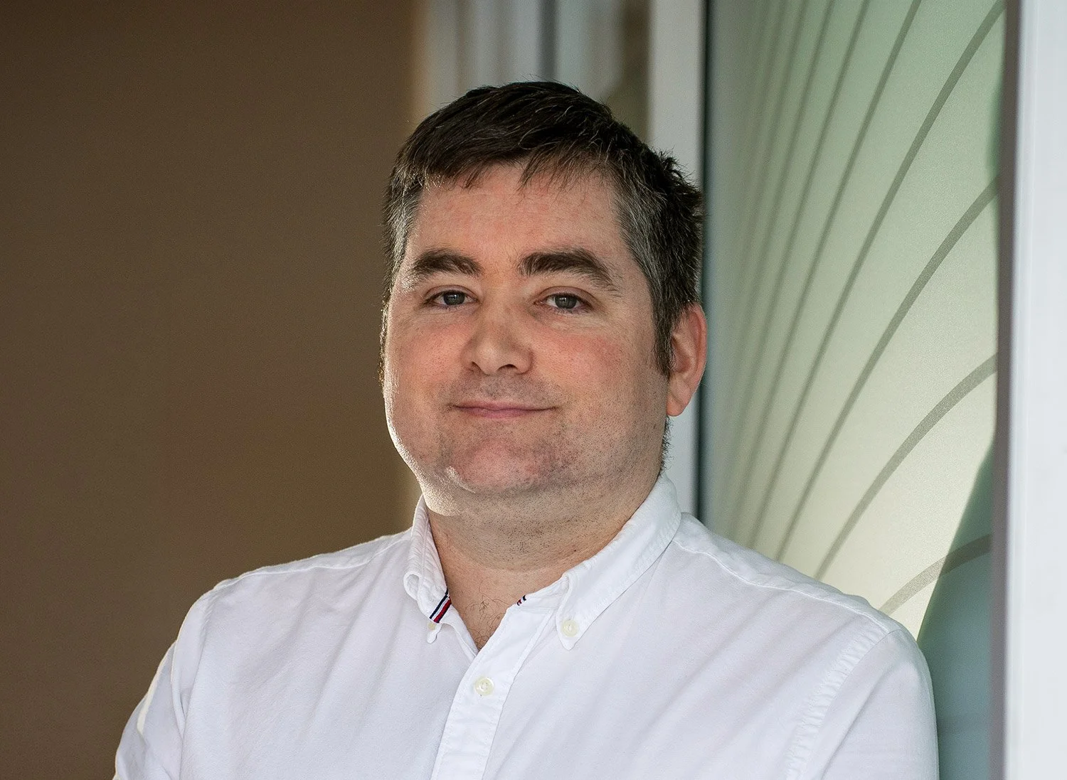 A man with dark hair, wearing a white collared shirt, standing near a window with a patterned green wall in the background.