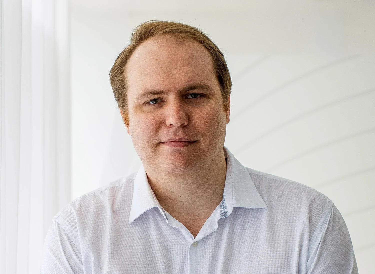 Portrait of a man with light brown hair in a white shirt, standing in front of a light-colored background.