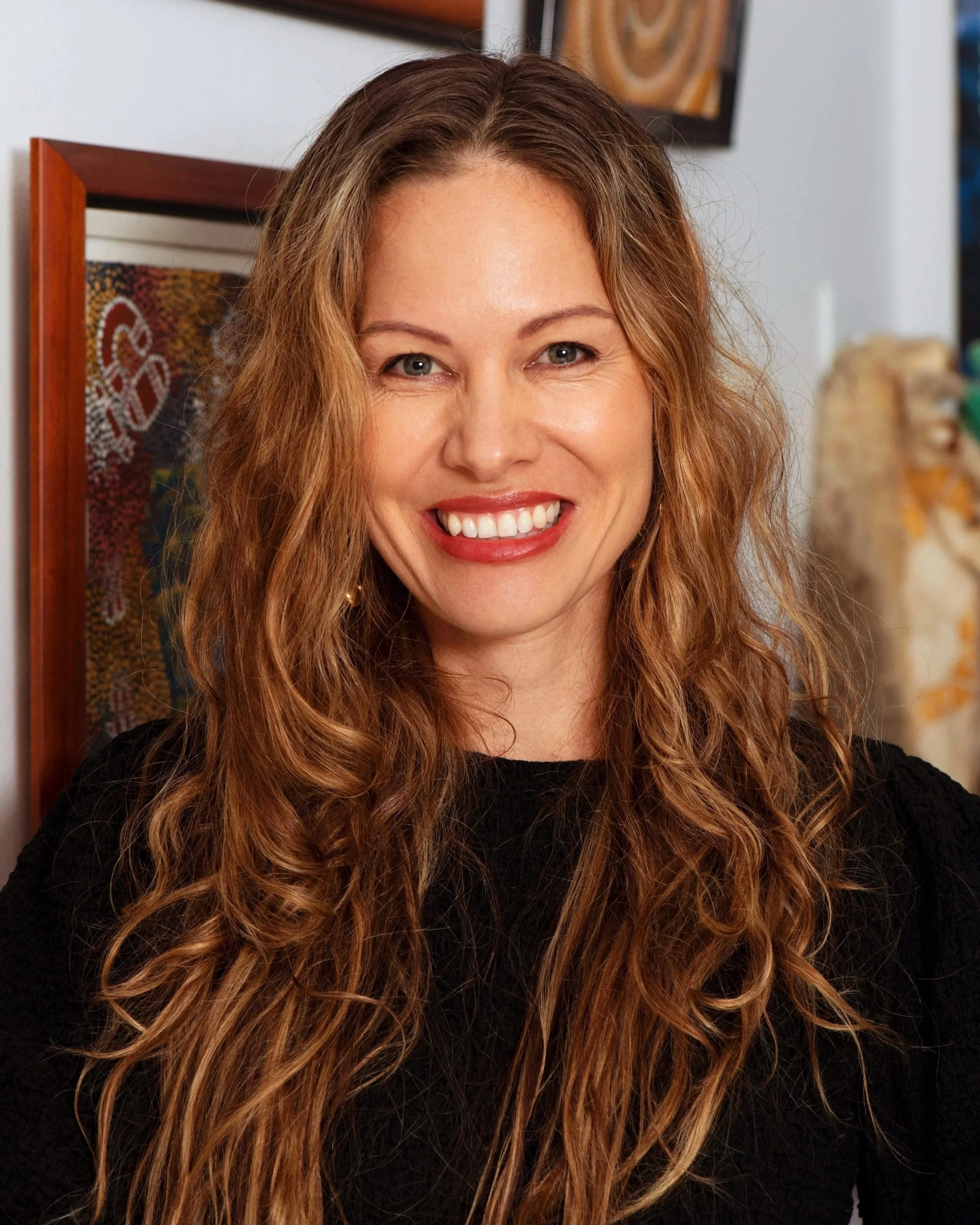 Samantha Laughton, Co-Managing Director at Honey Any Productions and a writer/director/producer.  A woman with long, wavy hair smiling at the camera, wearing a black top, with Indigenous artwork in the background.
