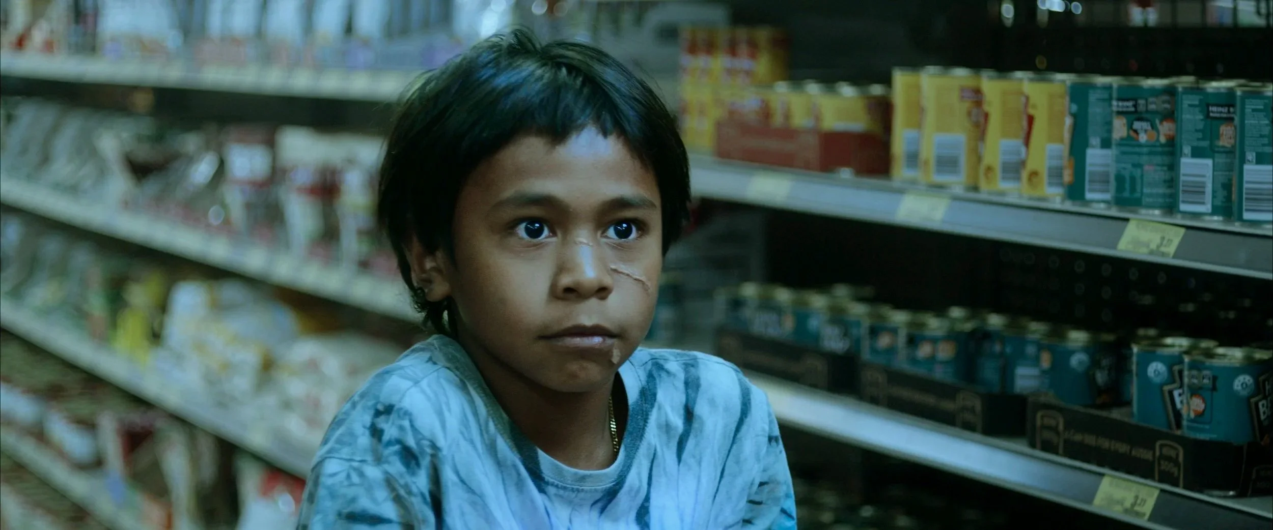 A young boy with a scrape on his face sitting in a grocery store aisle with shelves stocked with different packaged food products. Still from the short film 'Bear', by Rebecca Parker.