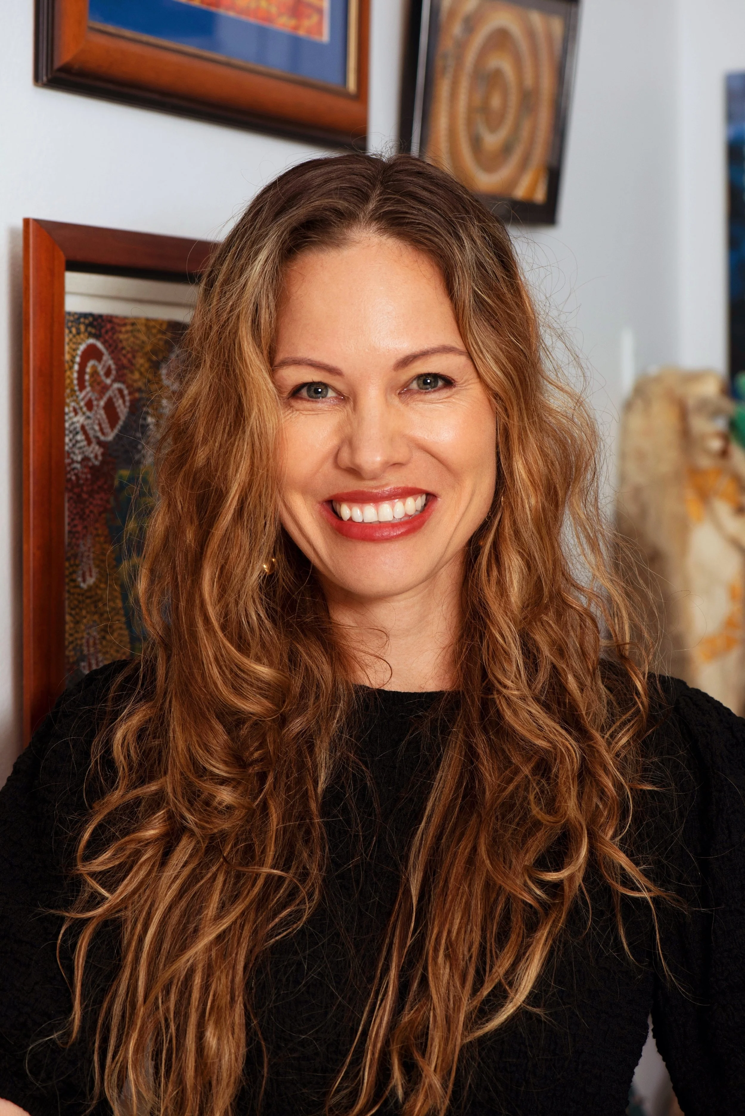 Samantha Laughton, Co-Managing Director at Honey Any Productions and a writer/director/producer.  A woman with long, wavy hair smiling at the camera, wearing a black top, with Indigenous artwork in the background.