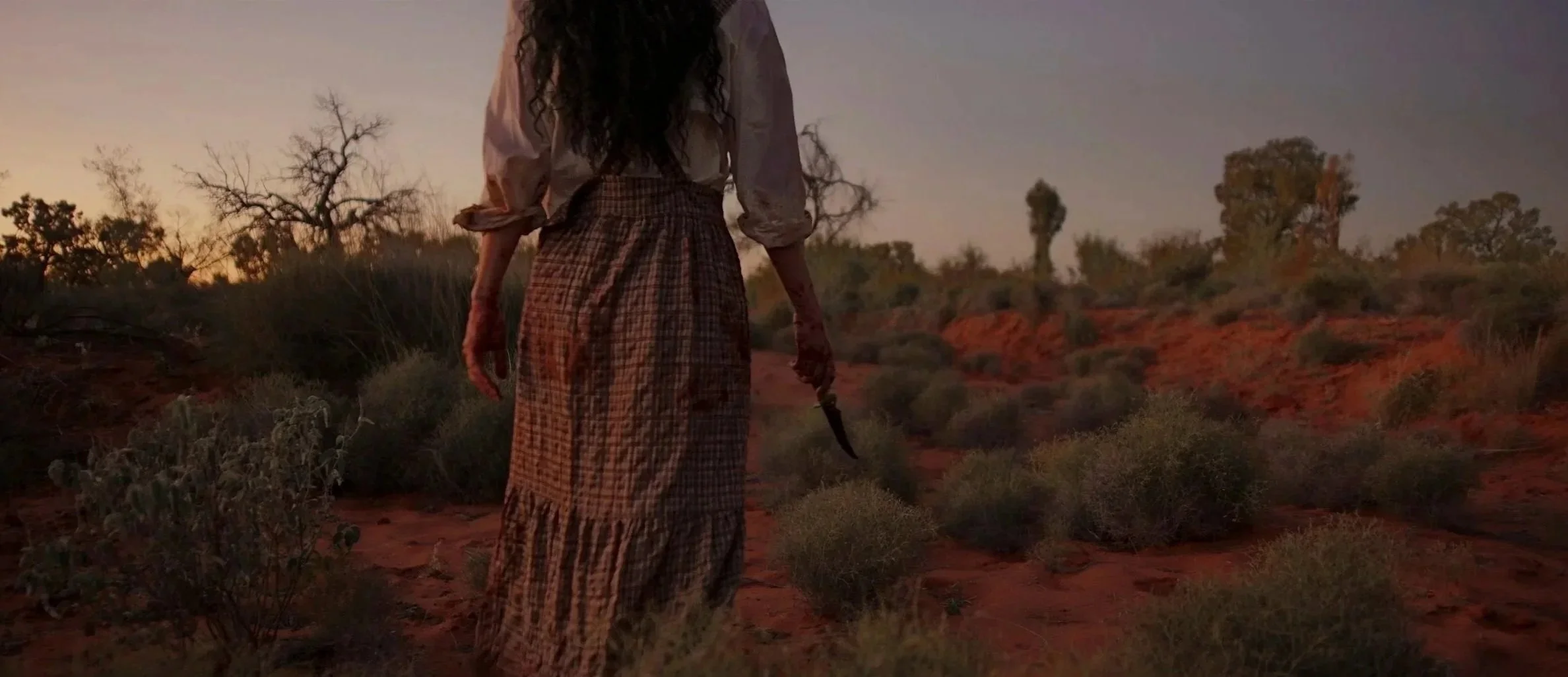 A woman in a long plaid skirt and loose blouse standing in a desert landscape during sunset, holding a knife in her right hand.