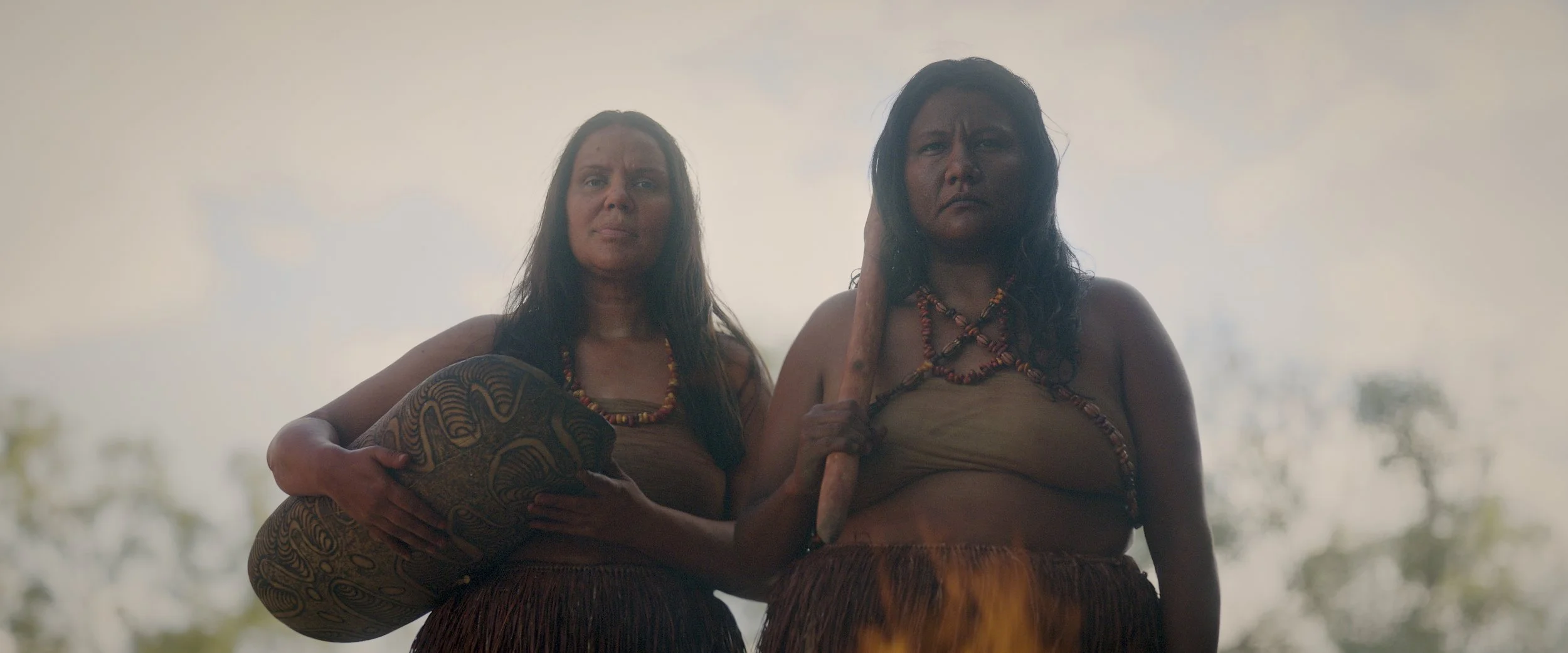 Two indigenous women in traditional attire, holding a carved wooden object, standing outdoors with a cloudy sky in the background.