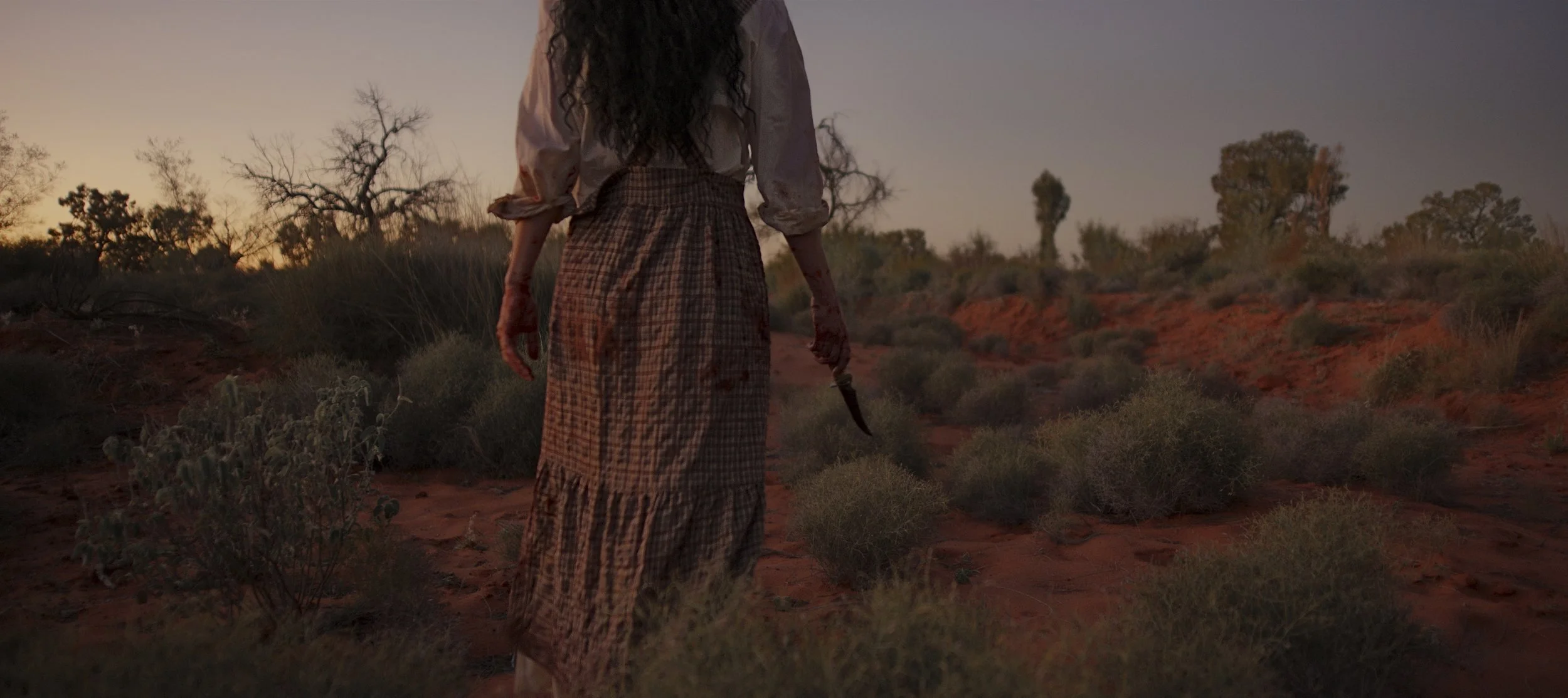 An Indigenous woman with long curly hair walking through a desert landscape at sunset, holding a knife in her right hand. Still image from the short film 'Fence' by Samantha Alexis Laughton.