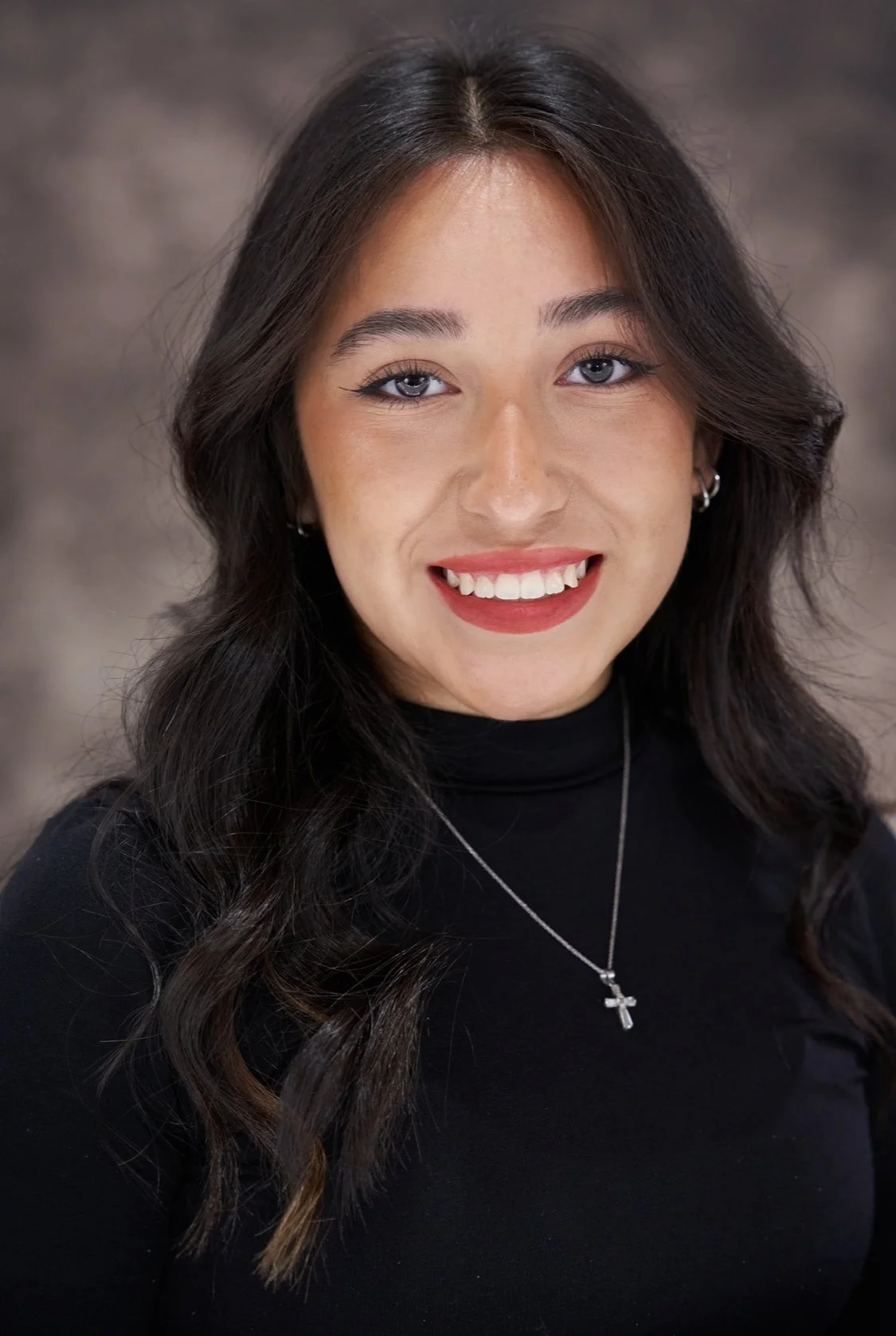 A young woman with dark wavy hair, wearing a black turtleneck and silver cross necklace, smiling in front of a blurred background.