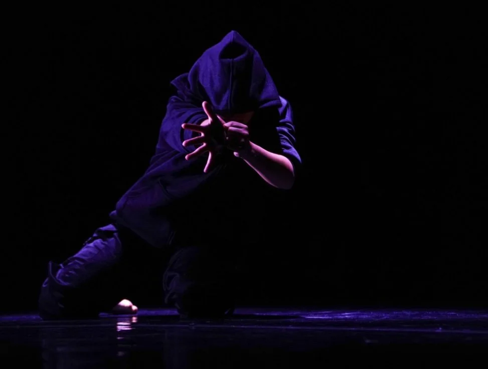 Person in dark hoodie performing dance move under purple stage lights, crouching on stage with hands extended