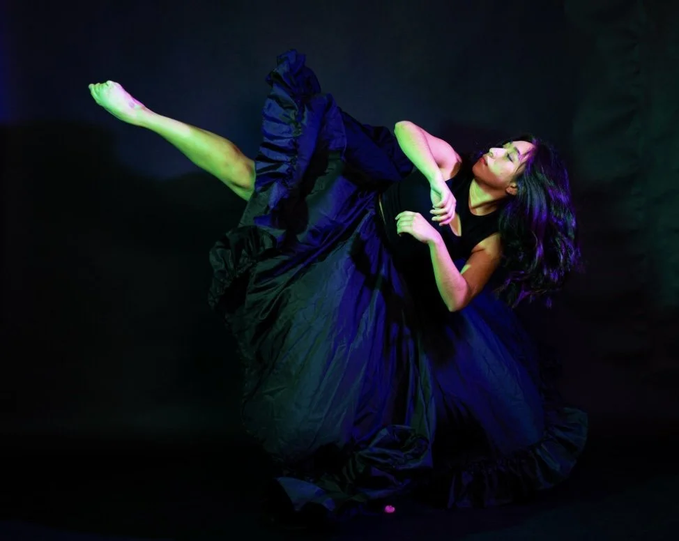 A woman with long dark hair wearing a black dress, in a dance pose against a black background with colorful lighting.