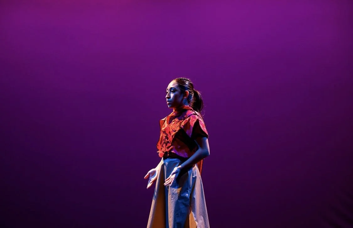 A woman standing alone on a stage illuminated with purple and blue lights, wearing a red floral top and beige wide-legged pants.
