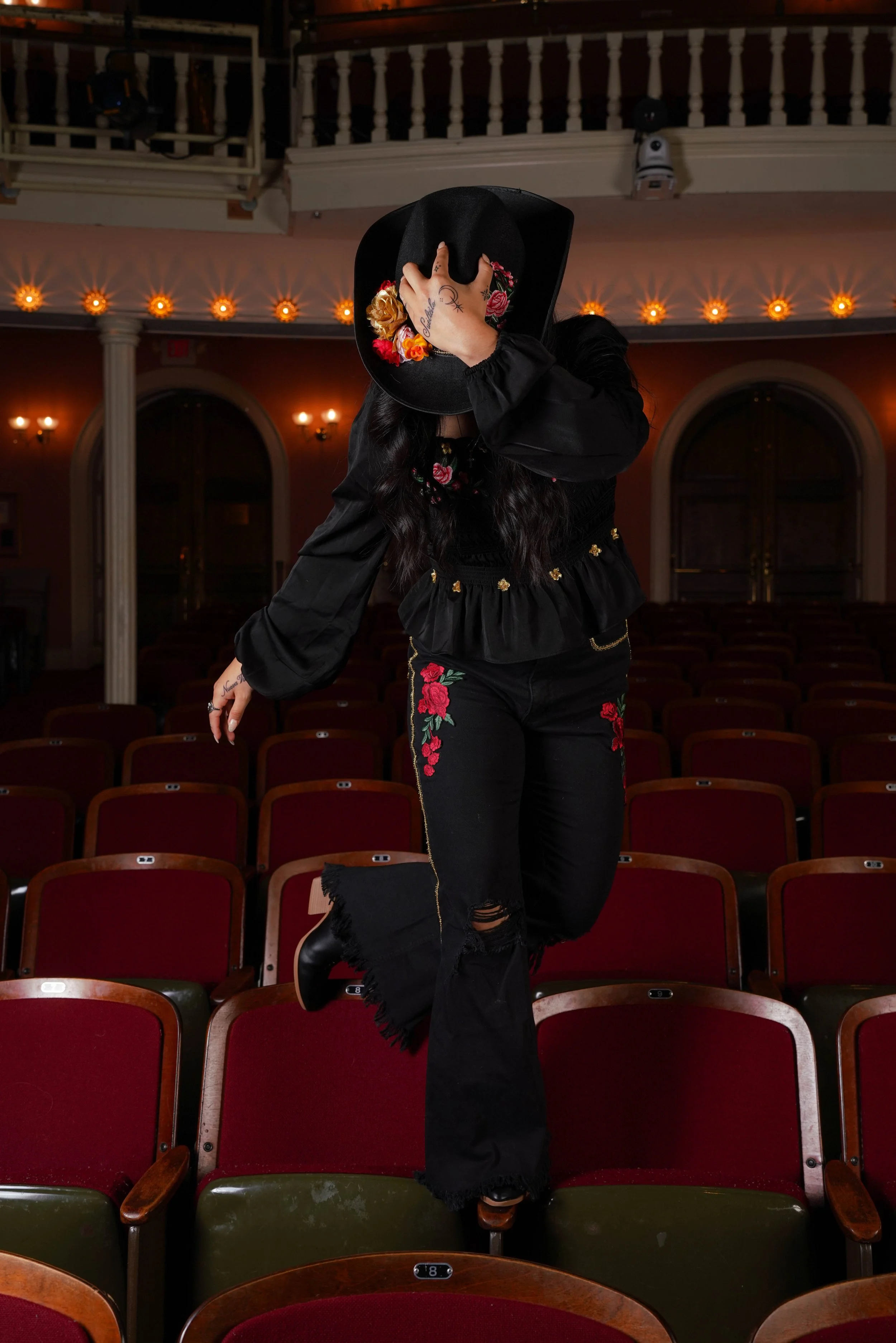 A woman dressed in black with floral embroidery, wearing a large hat adorned with flowers, standing on theater seats inside a vintage style theater or auditorium with red velvet seats and ornate lighting fixtures.