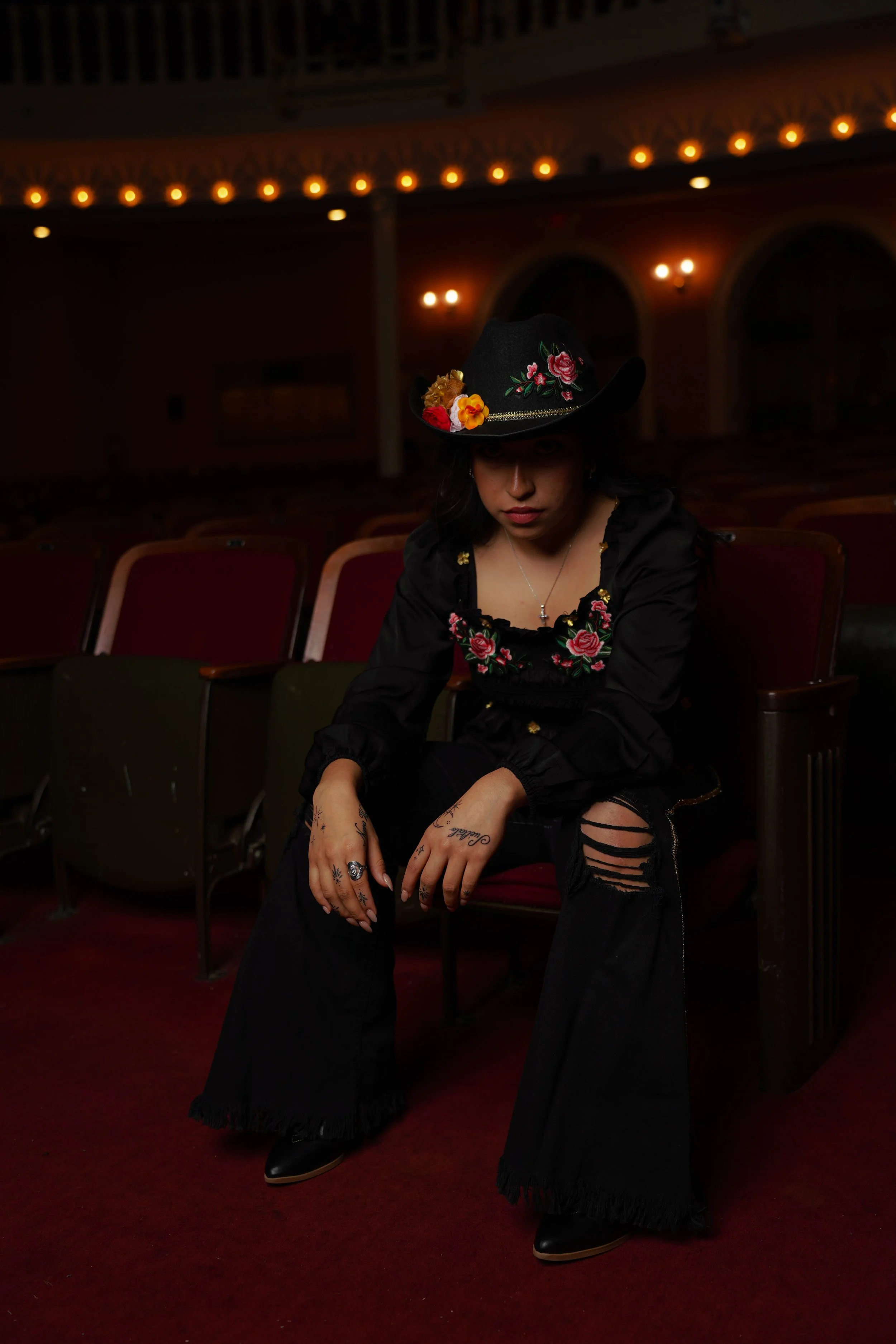 A young woman sits alone in an auditorium with red carpet and empty red seats. She wears a black hat with colorful embroidered flowers, a black embroidered top, ripped black jeans, and black boots. She has tattoos on her hands and rings.