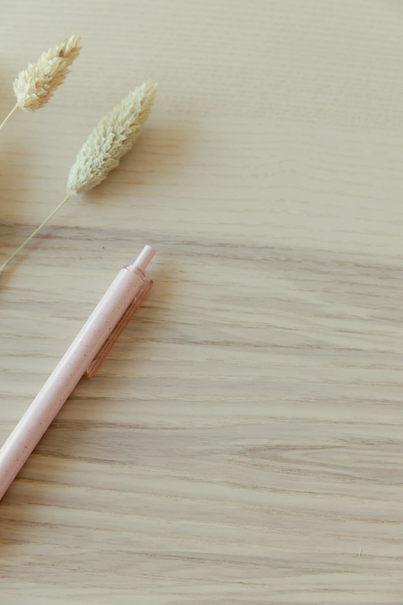 Two beige dried wheat stalks and a light pink cream tube lying on a wooden surface.